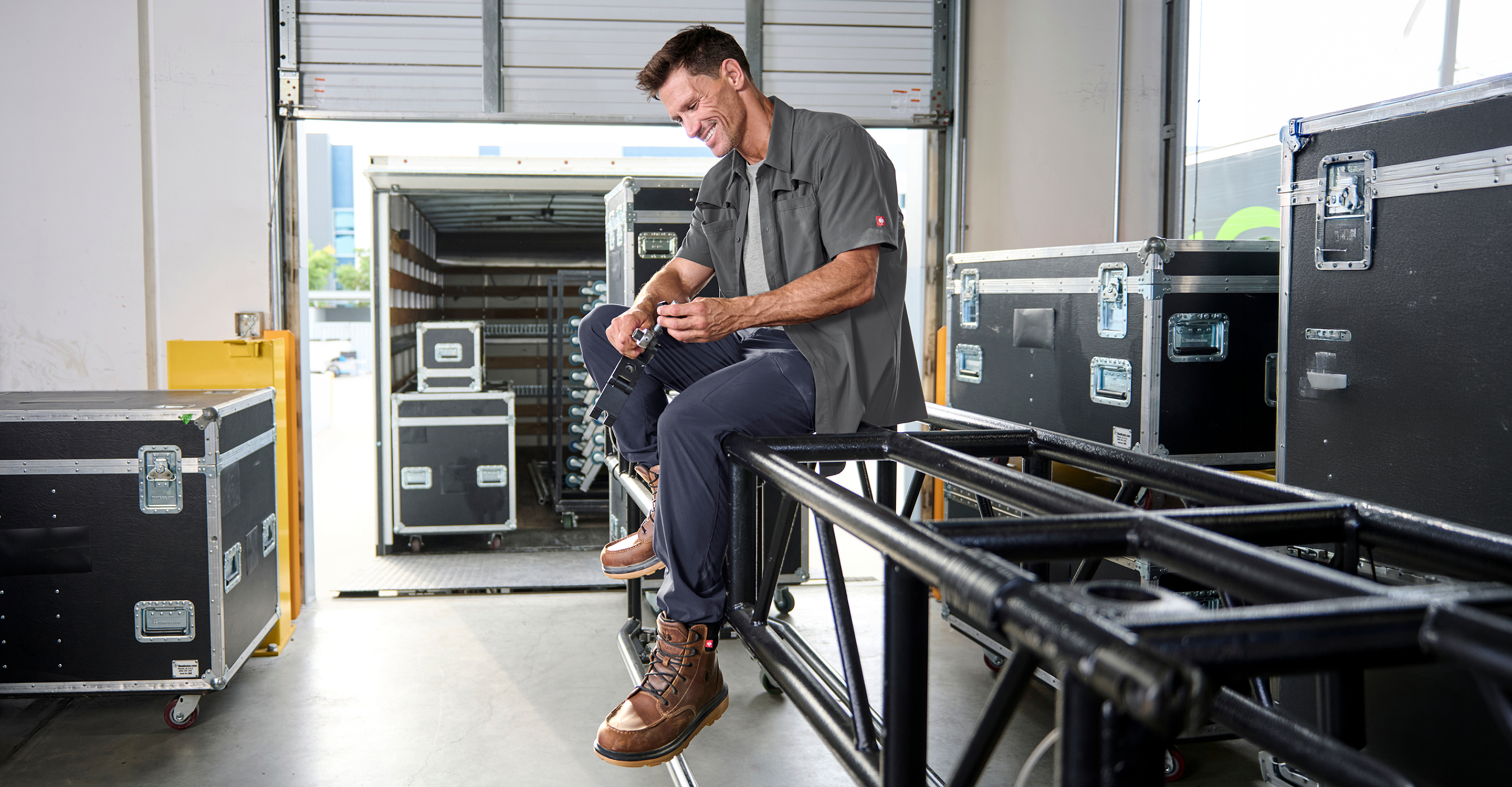 A man in work boots and a gray shirt sits on metal pipes, smiling, with equipment cases and an open truck in a warehouse setting.