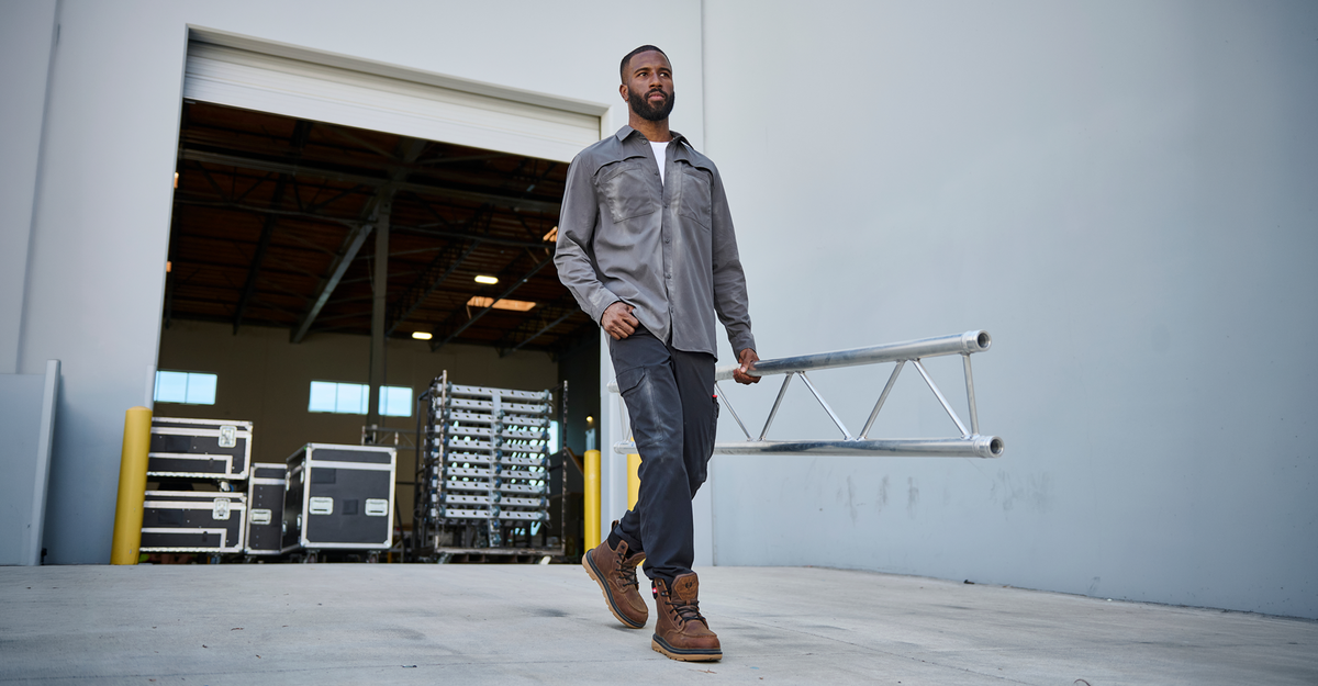 A man in a gray work shirt and dark pants walks through a warehouse doorway while carrying a silver metal truss bar.