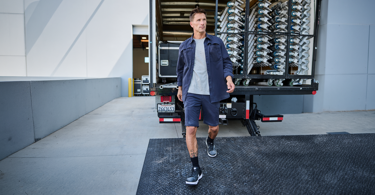 A man in a blue work jacket and shorts walks away from a truck filled with organized packages or containers, outside a modern building on a sunny day.