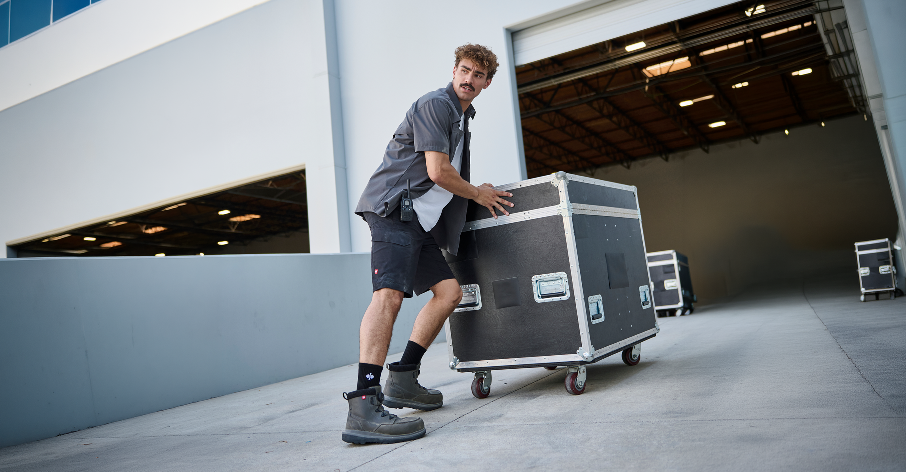 A man in work clothes and boots pushes a large black equipment case on wheels outside a warehouse loading dock.
