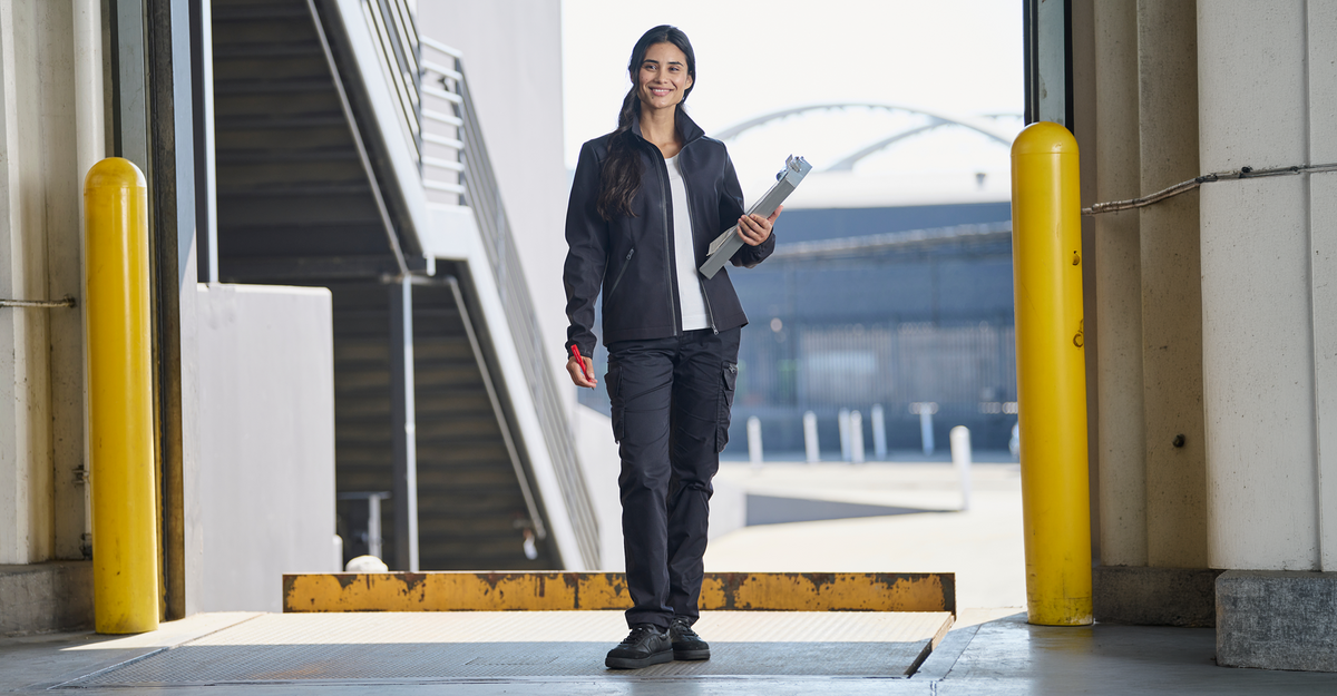 A woman in work attire stands in an industrial setting, holding rolled-up papers and smiling. Yellow safety bollards and an outdoor staircase are visible in the background.