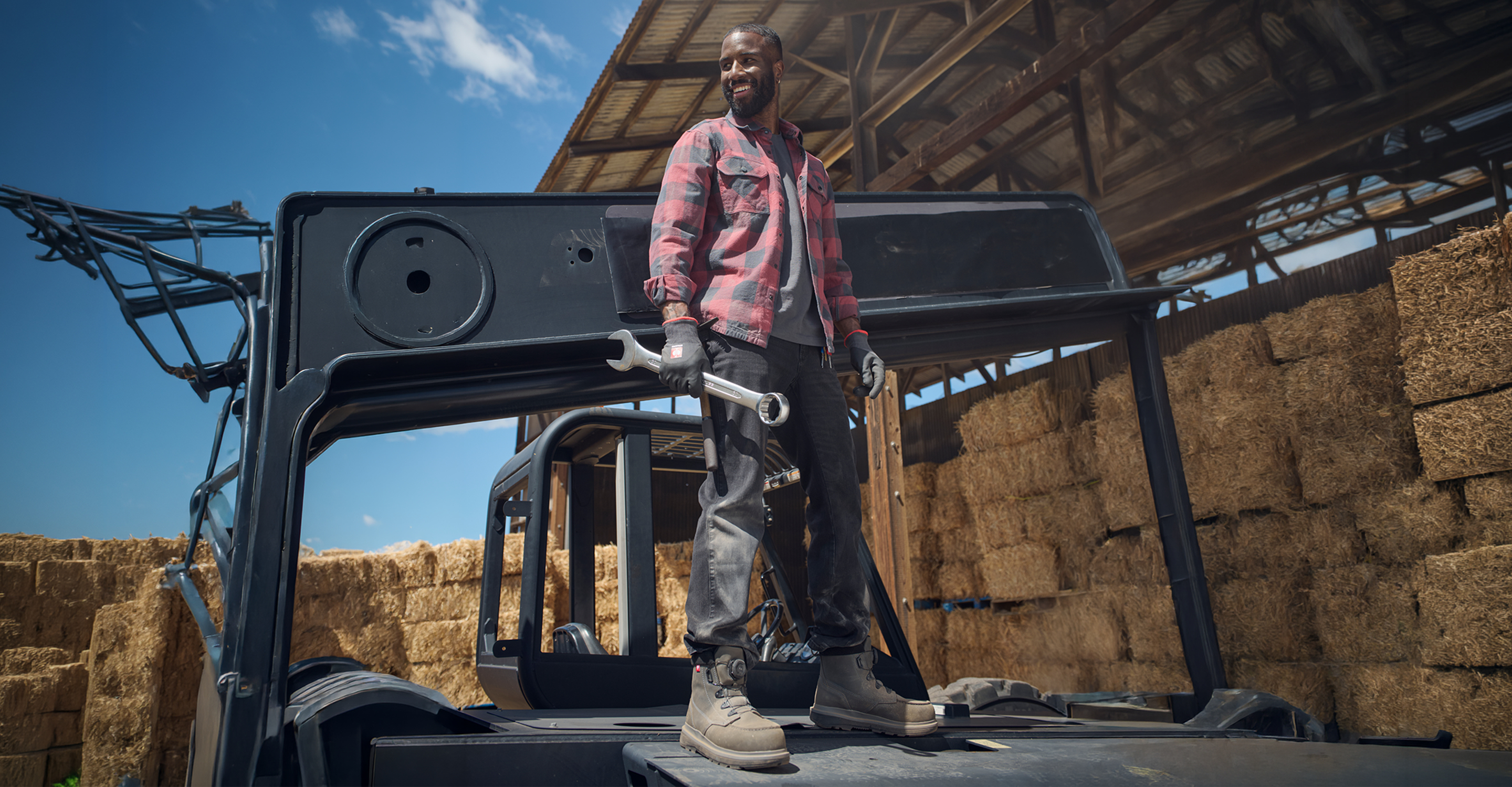 A man wearing a red and black plaid flannel shirt, dark gray work pants, and work boots stands on the cab of a large black agricultural tractor or loader in a barn with stacked hay bales under a blue sky.