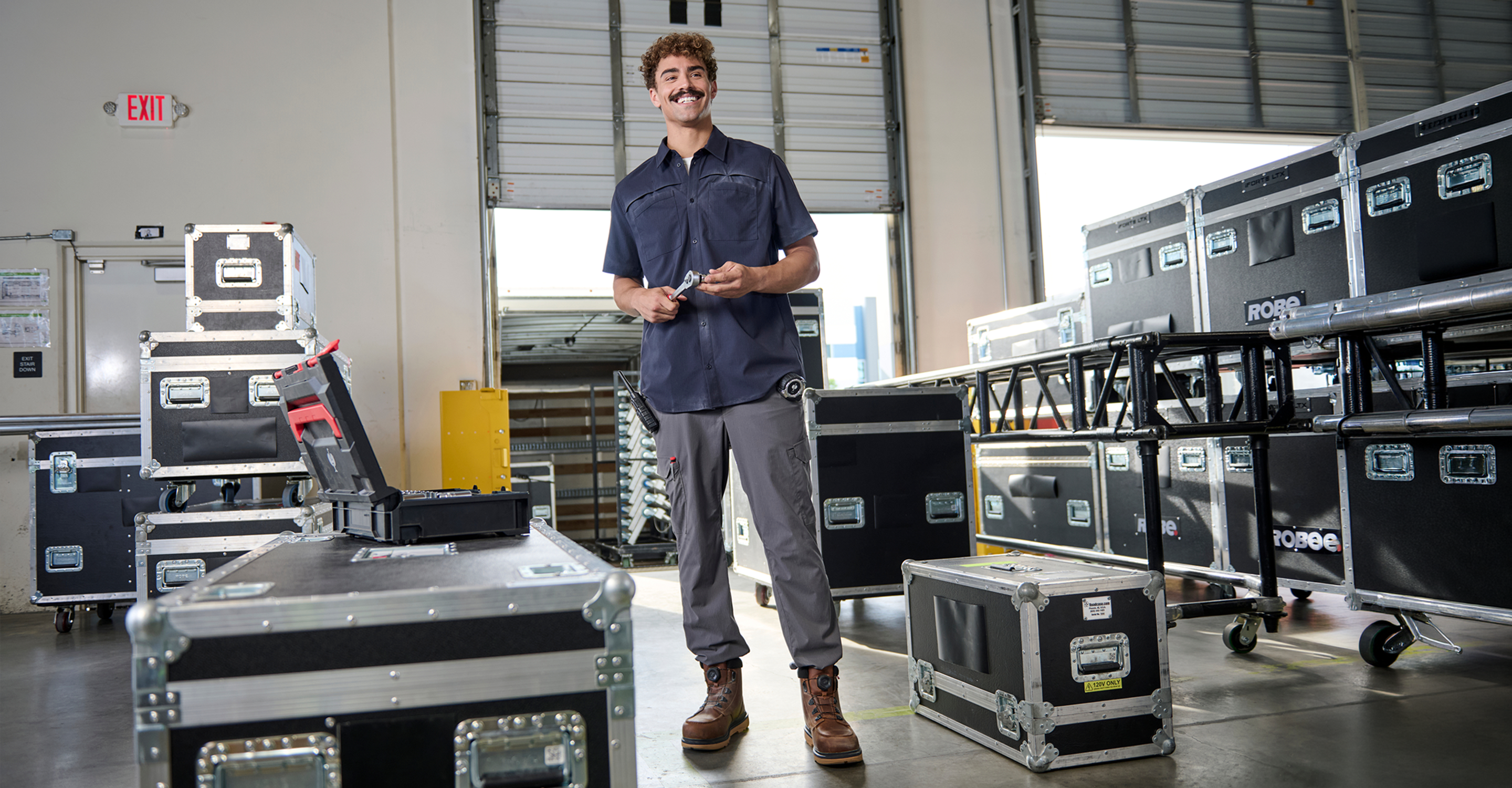 A man in work clothes stands in a warehouse surrounded by large black equipment cases, holding tools and smiling. The warehouse doors are open, letting in natural light.
