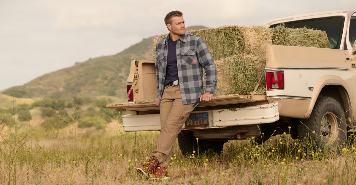 A man in a plaid flannel shirt and tan pants leans against the tailgate of a vintage pickup truck loaded with hay in an outdoor field.