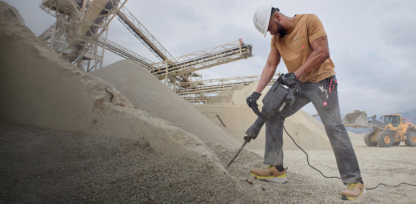 Worker operating a drill in an industrial setting with machinery and equipment in the background.