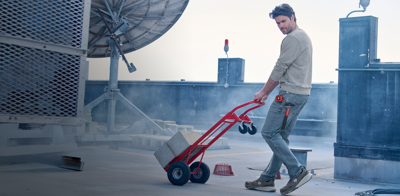 Man pushing a red hand truck on a rooftop with large satellite dish and equipment in the background.