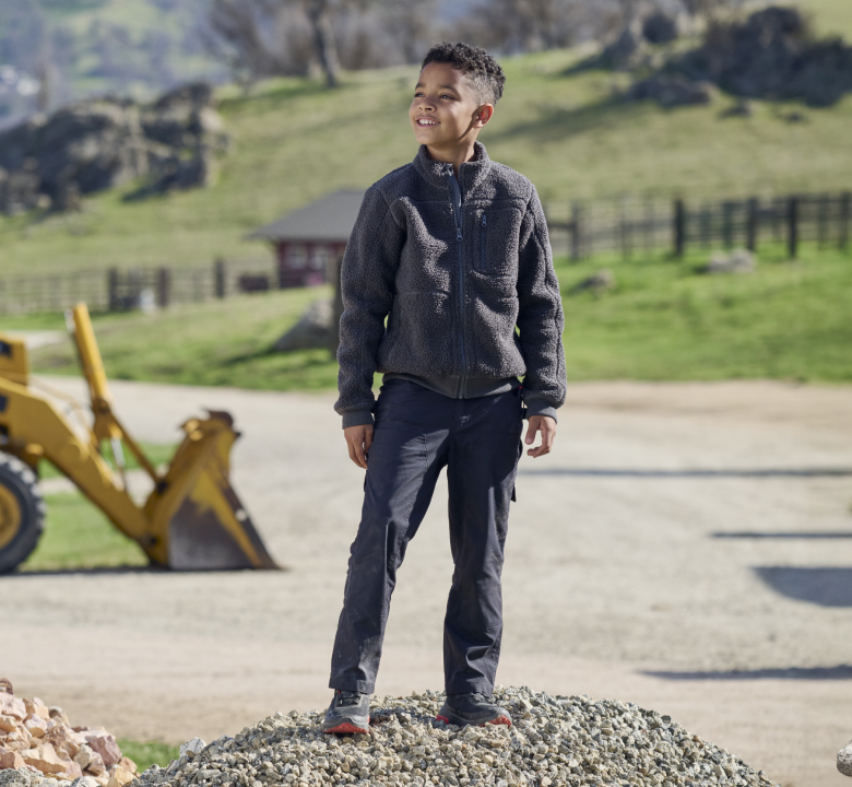 A smiling boy stands on a gravel mound outdoors, wearing a dark jacket and pants. A yellow backhoe and green fields with fences and trees are visible in the background.