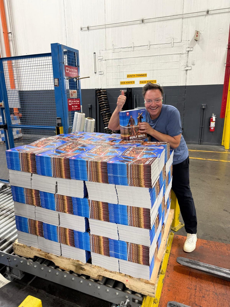 A smiling man in a warehouse stands behind a large pallet stacked with printed book or box packages wrapped in blue and white stretch bands. He holds a small printed item above the pallet, giving a thumbs-up in a bright industrial setting with shelving and equipment visible.