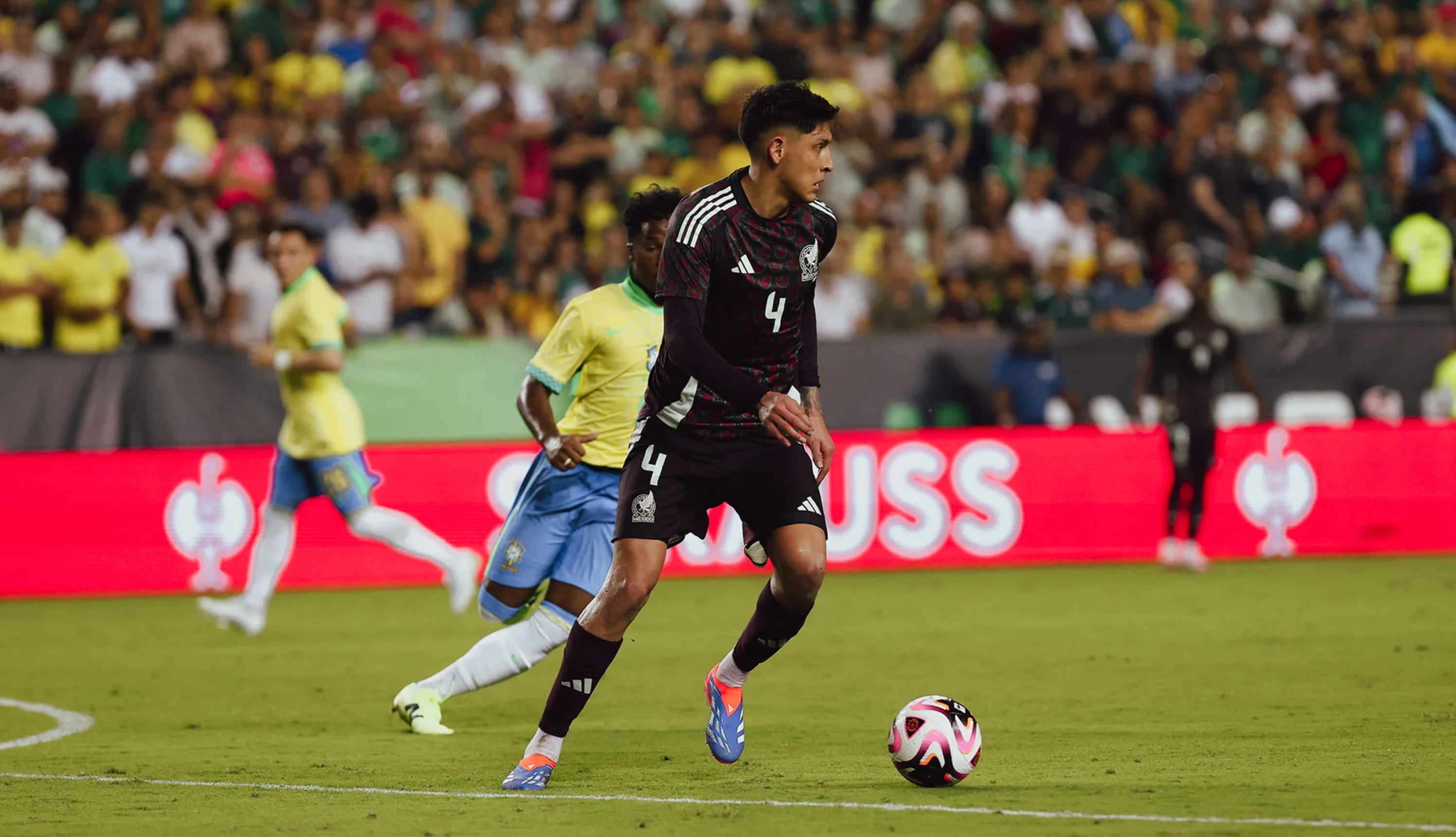 Soccer player in a black uniform, number 4, dribbles a pink and white ball on a field during a match, with opponents in yellow and blue and a crowd in the background.