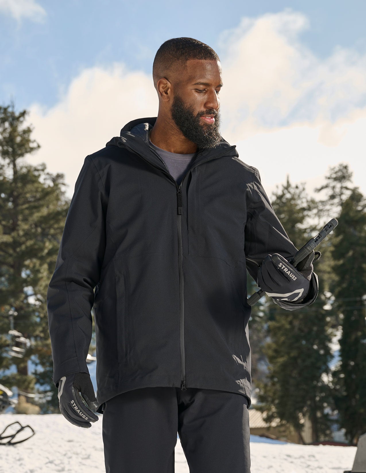 Man wearing a black jacket and gloves standing outdoors with trees and sky in the background.