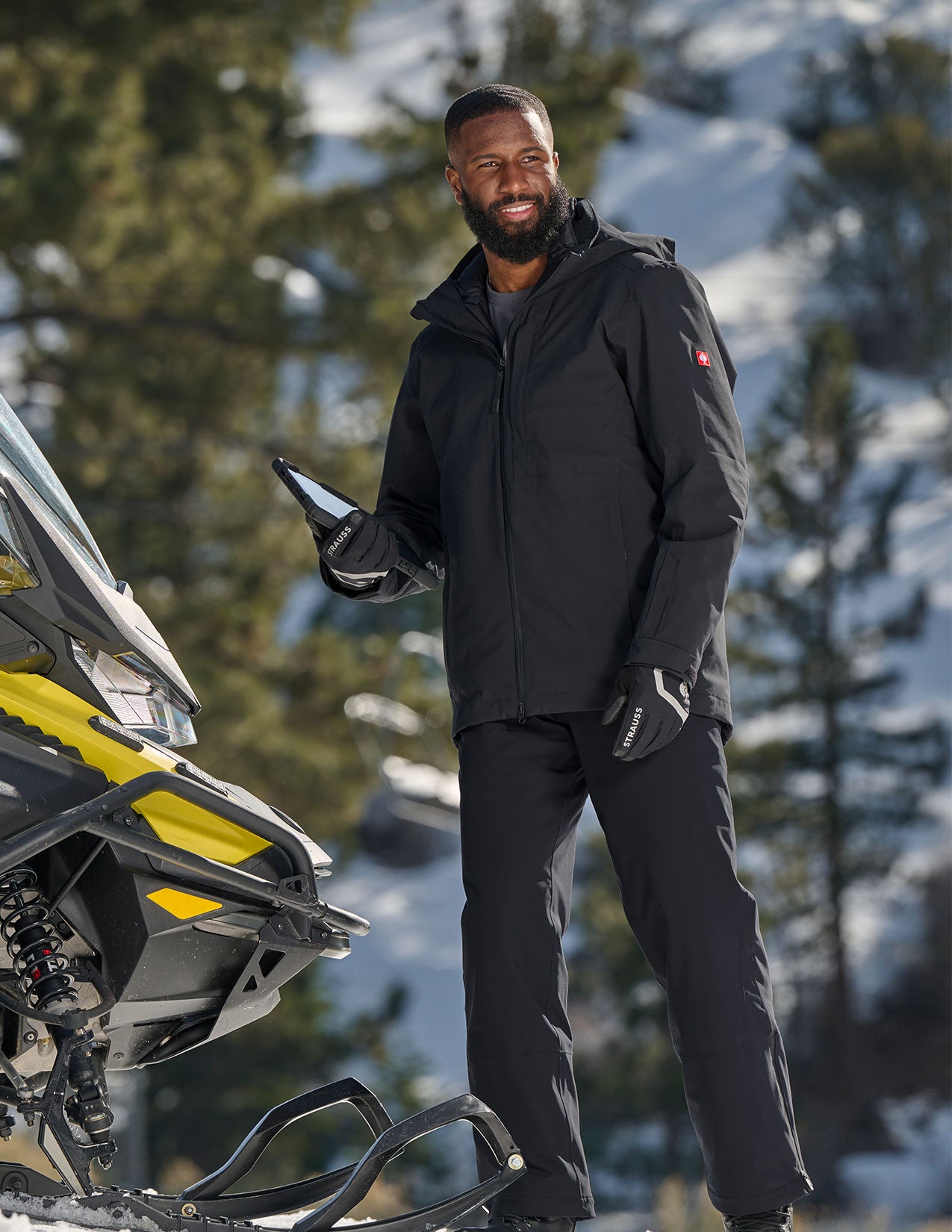 Man in black winter clothing standing next to a snowmobile in a snowy forest.