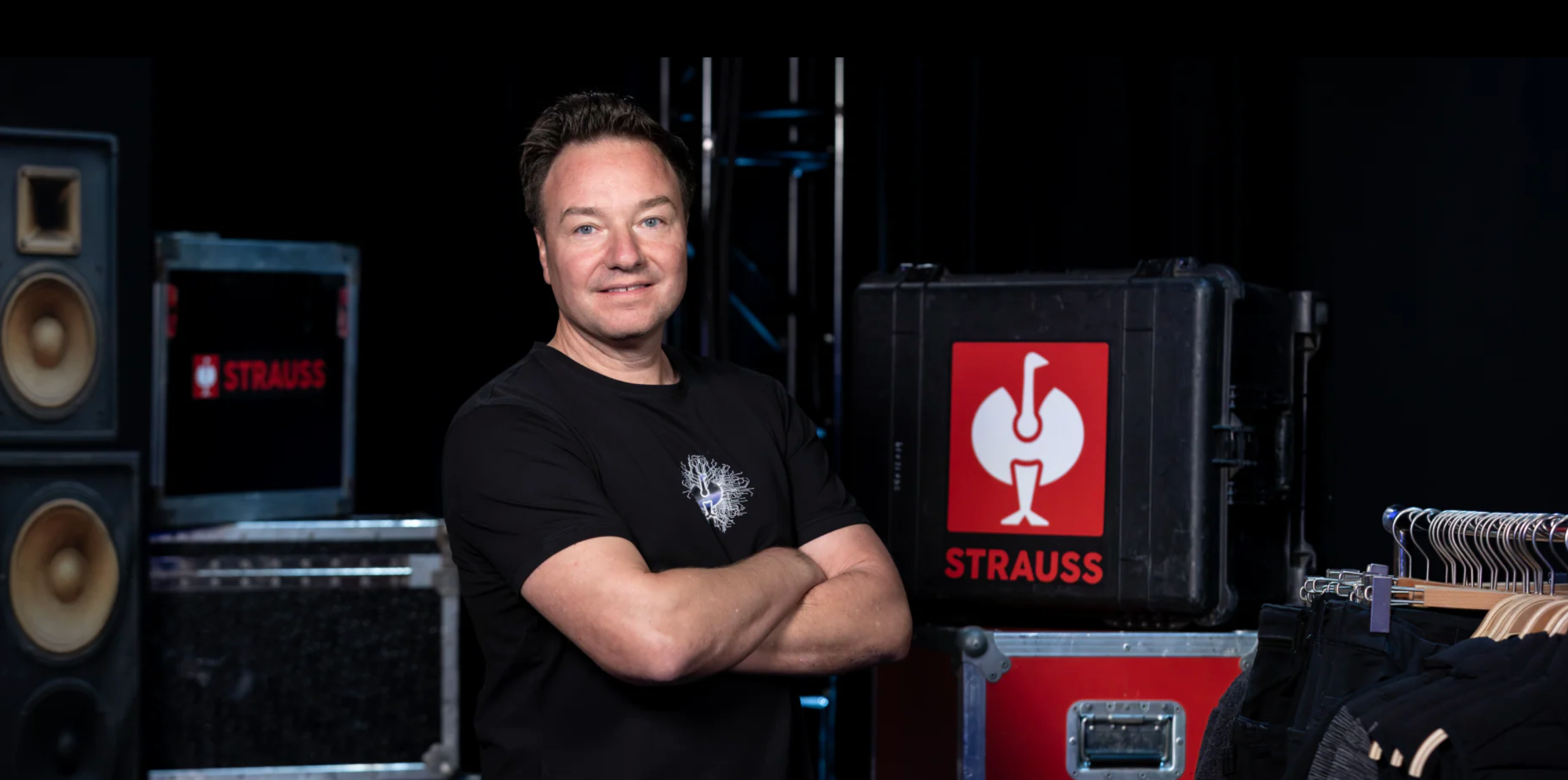 A man with folded arms stands in front of equipment cases and a clothing rack, with a red Strauss logo featuring a white bird visible on one of the cases.