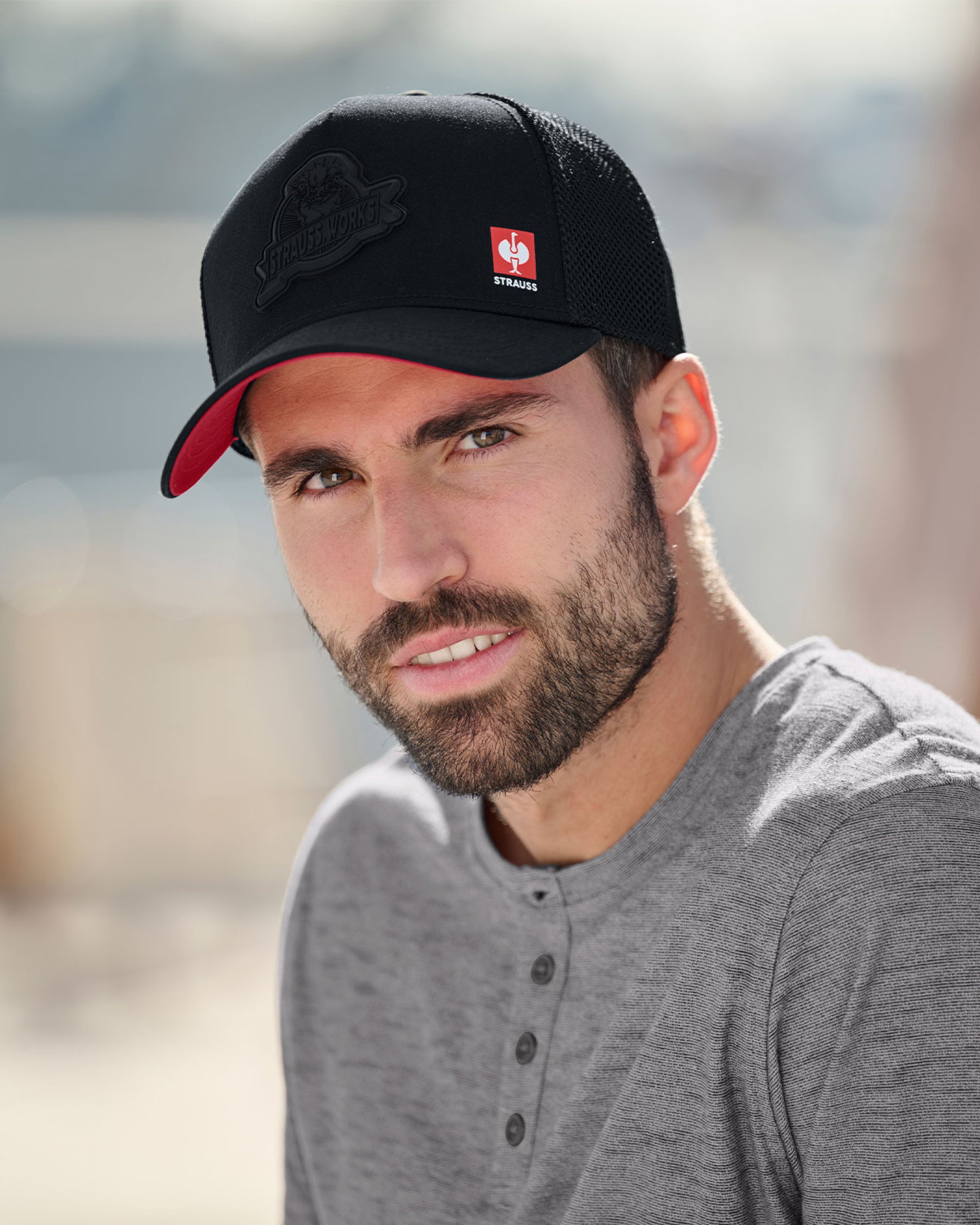 A worker wears a black STRAUSS Workwear trucker hat with a work shirt and utility pants outdoors.