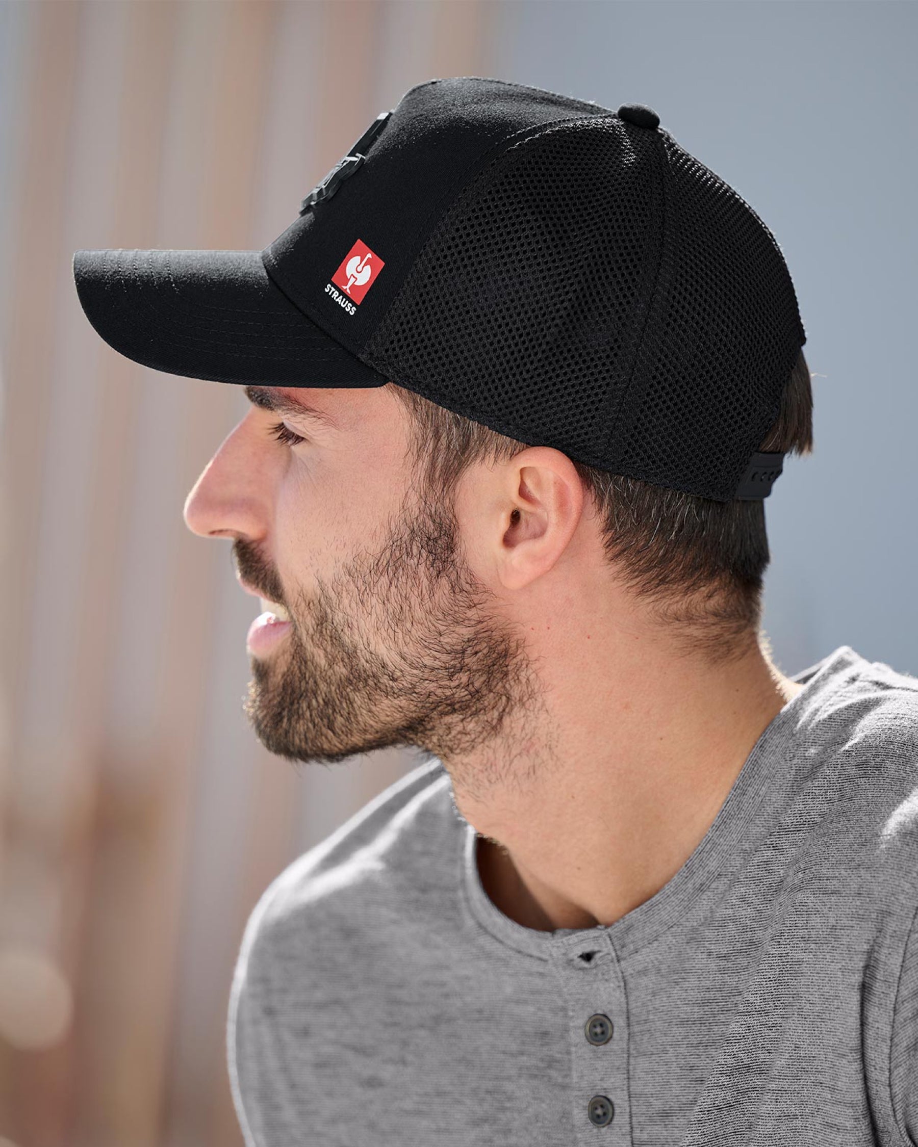 A worker in a black STRAUSS Workwear trucker hat stands near jobsite materials in outdoor workwear.