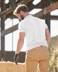 A worker wears the STRAUSS Performance Polo Shirt in white with tan work pants beside jobsite equipment