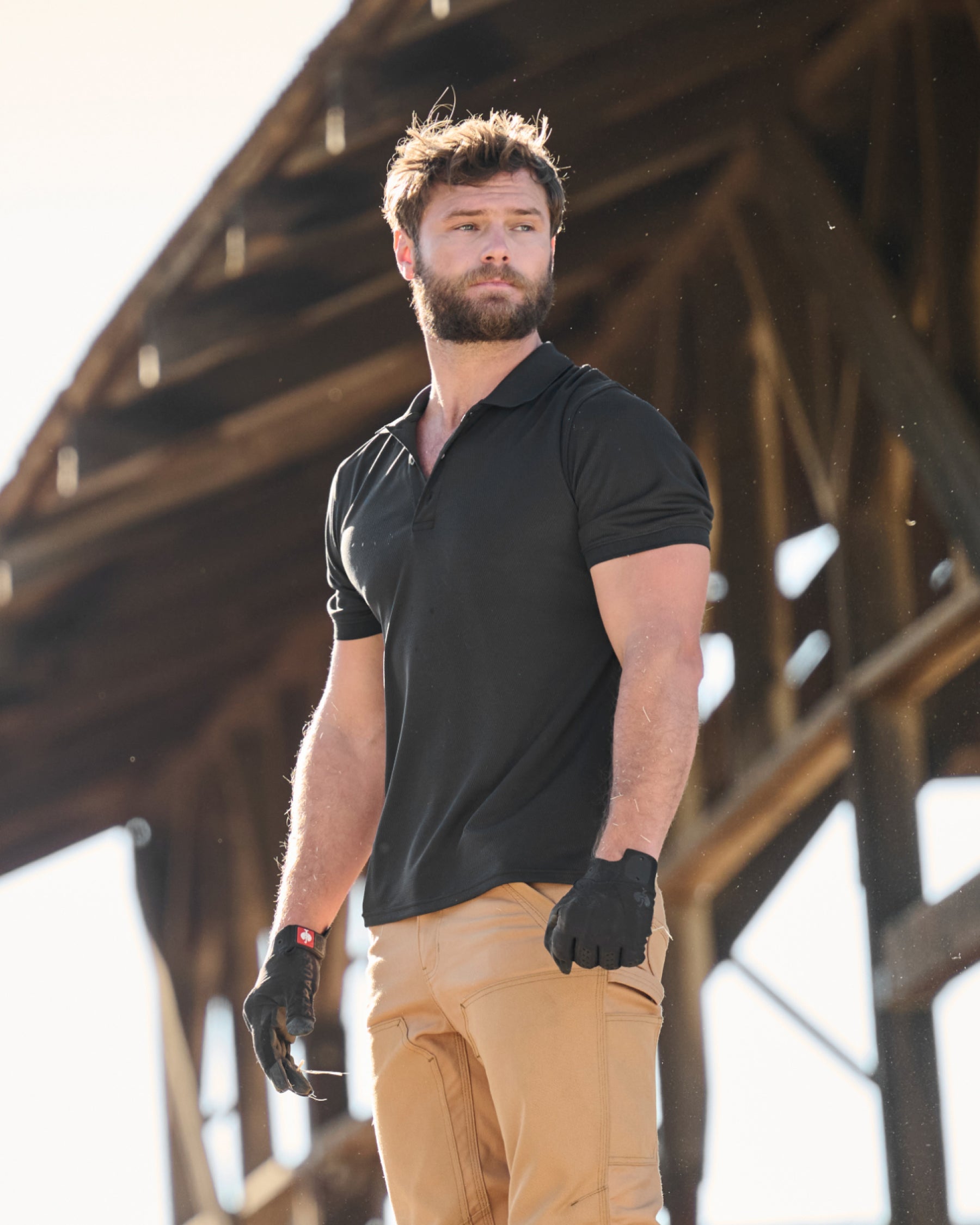 A worker wears the STRAUSS Performance Polo Shirt in black with tan work pants on a sunny jobsite