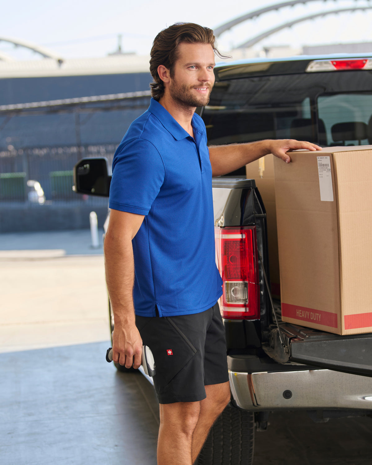 A worker wears the STRAUSS Performance Polo Shirt in royal blue beside jobsite equipment