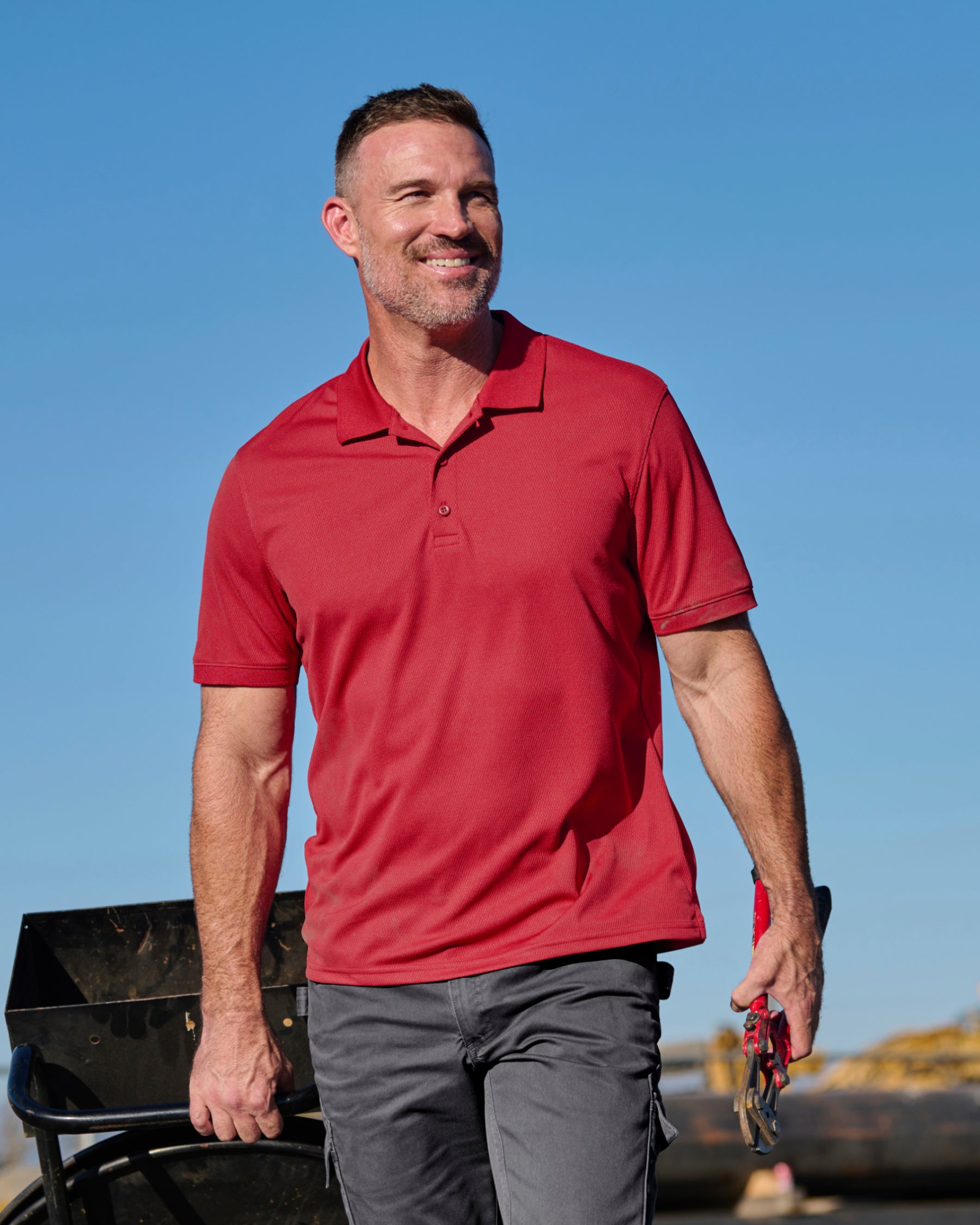 A worker wears the STRAUSS Performance Polo Shirt in fire red with tan work pants on a sunny jobsite