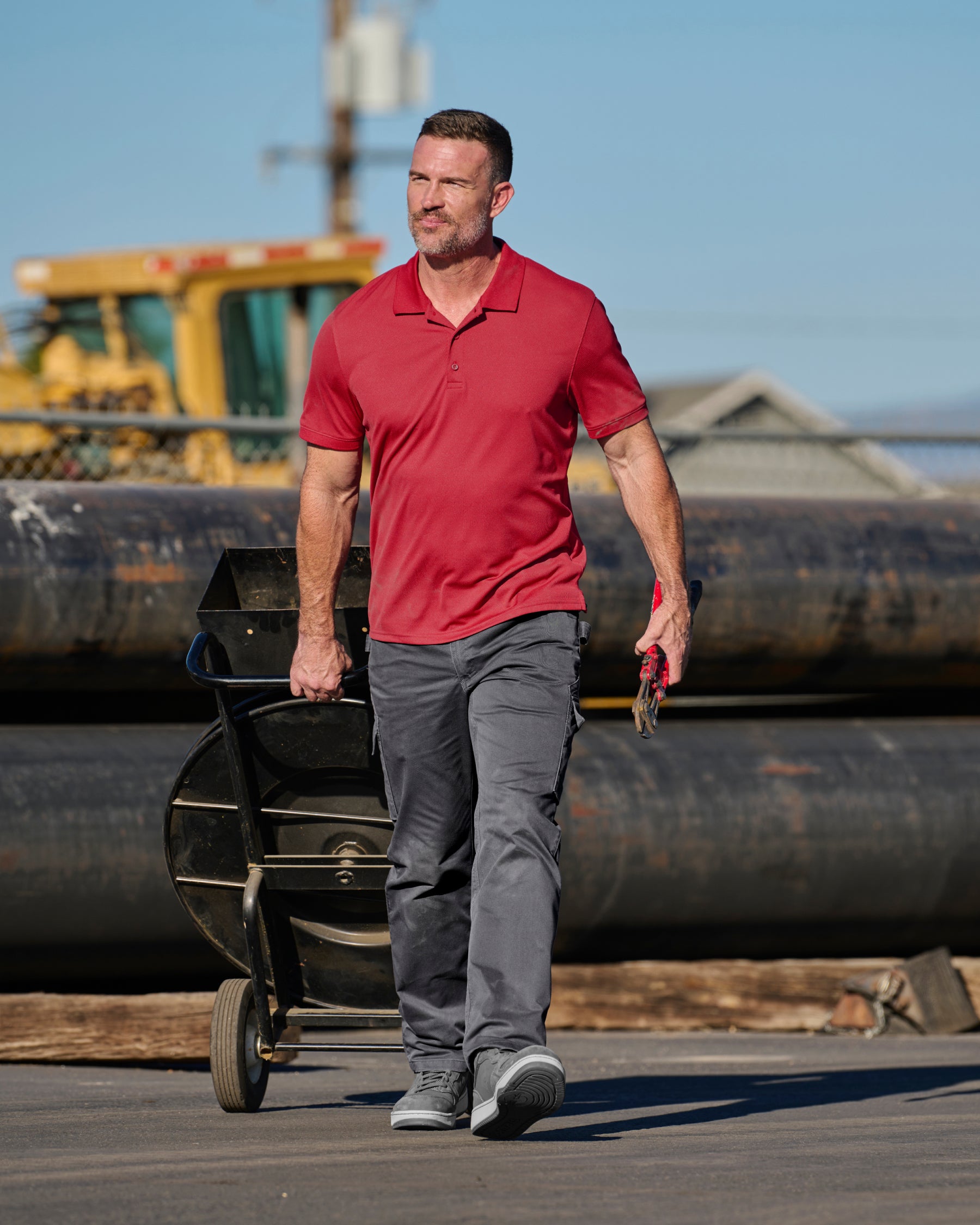 A worker wears the STRAUSS Performance Polo Shirt in fire red with tan work pants while working in a field