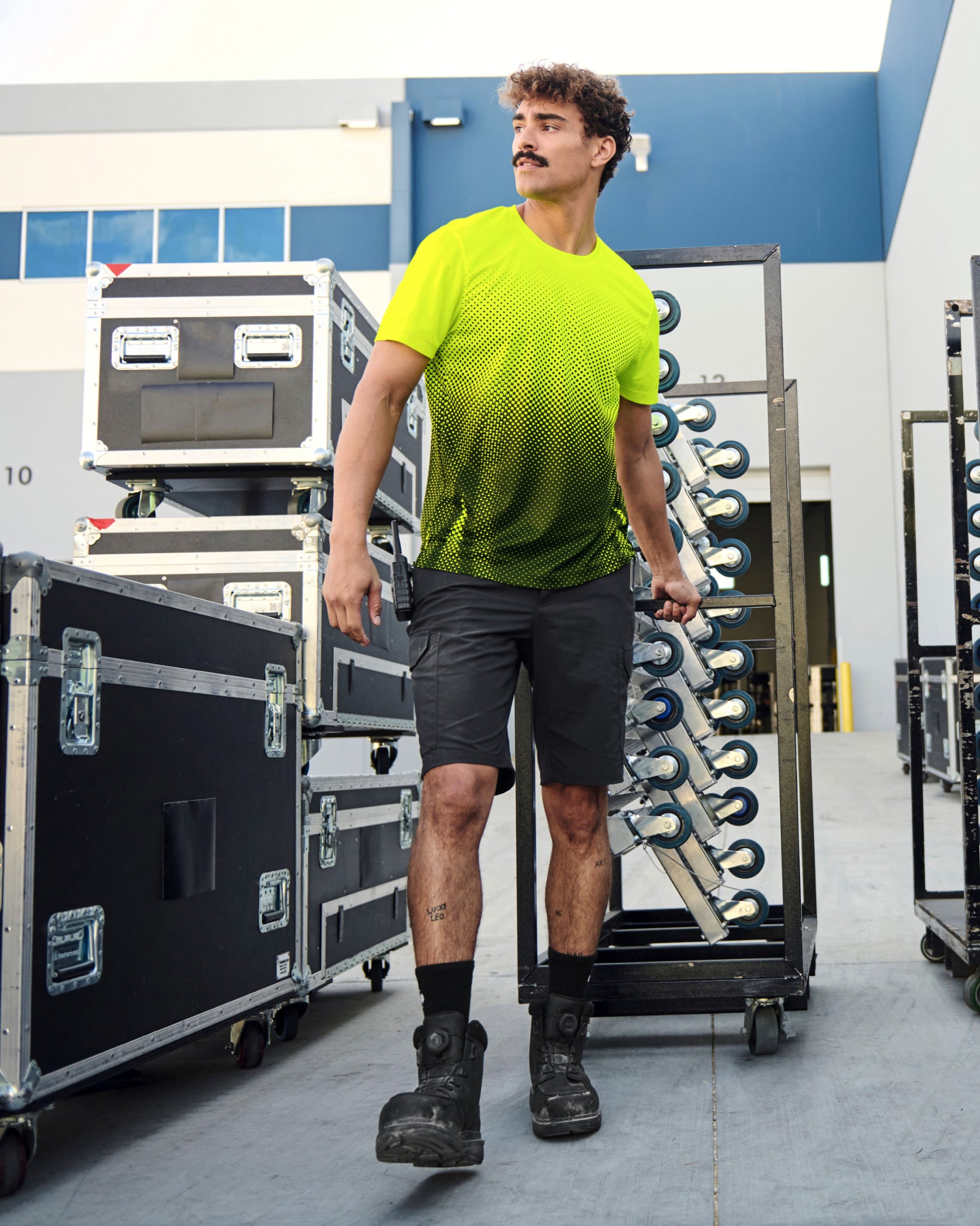 A worker wears the STRAUSS Enhanced Visibility Performance T-Shirt e.s.t:aktik in neon yellow and black with black work pants near equipment on a paved jobsite