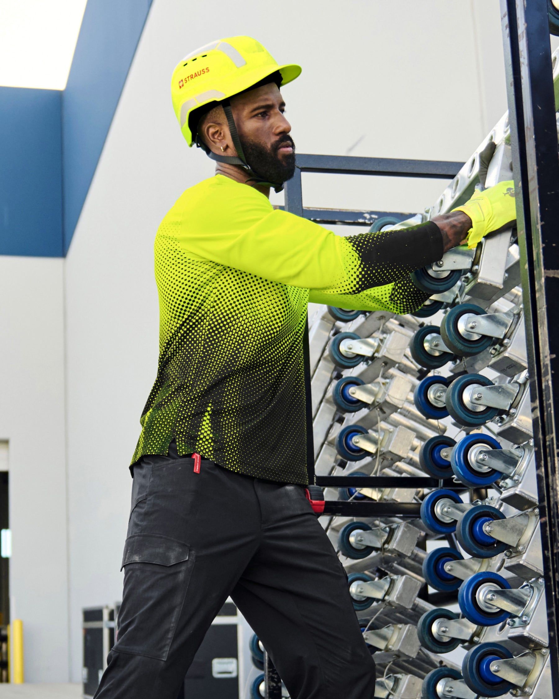 A worker wears the STRAUSS Enhanced Visibility Performance Long Sleeve Shirt e.s.t:aktik in neon yellow and black with black work pants beside construction fencing