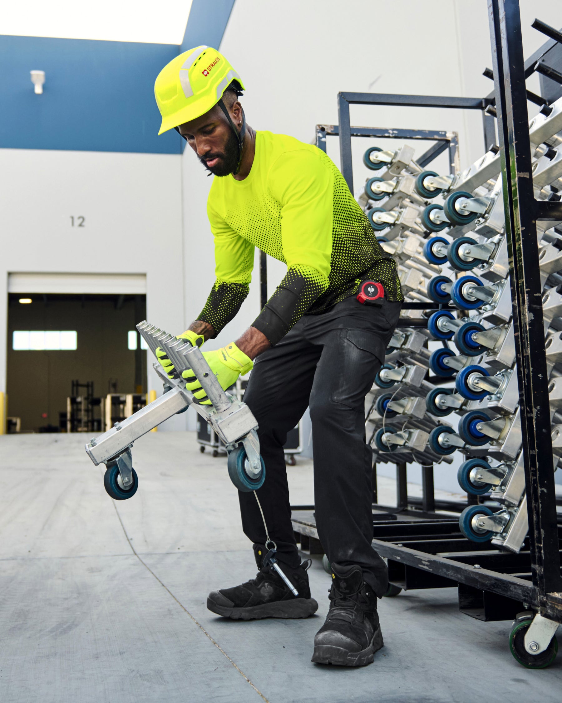 A worker wears the STRAUSS Enhanced Visibility Performance Long Sleeve Shirt e.s.t:aktik in neon yellow and black with black work pants near equipment on a paved jobsite