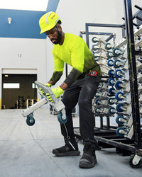 A worker wears the STRAUSS Enhanced Visibility Performance Long Sleeve Shirt e.s.t:aktik in neon yellow and black with black work pants near equipment on a paved jobsite