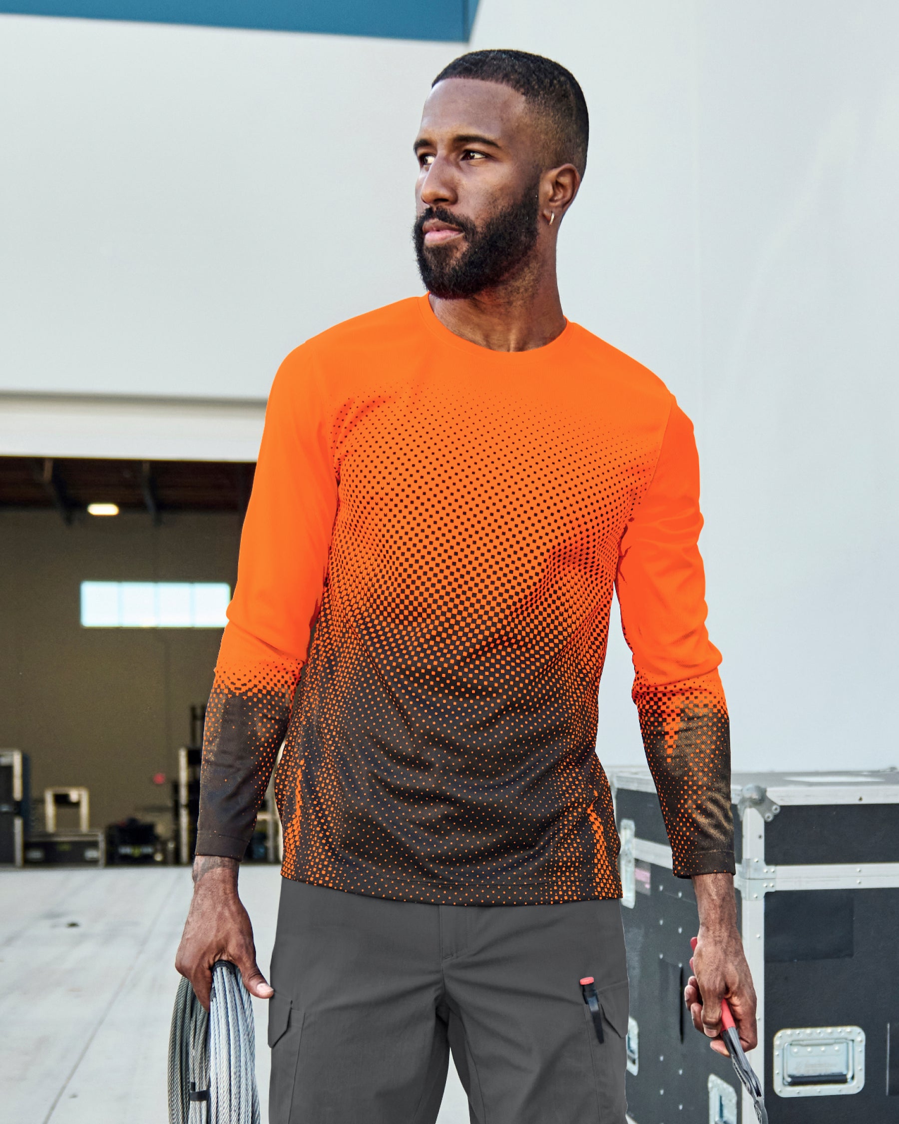 A worker wears the STRAUSS Enhanced Visibility Performance Long Sleeve Shirt e.s.t:aktik in neon orange and black with black work pants beside construction fencing