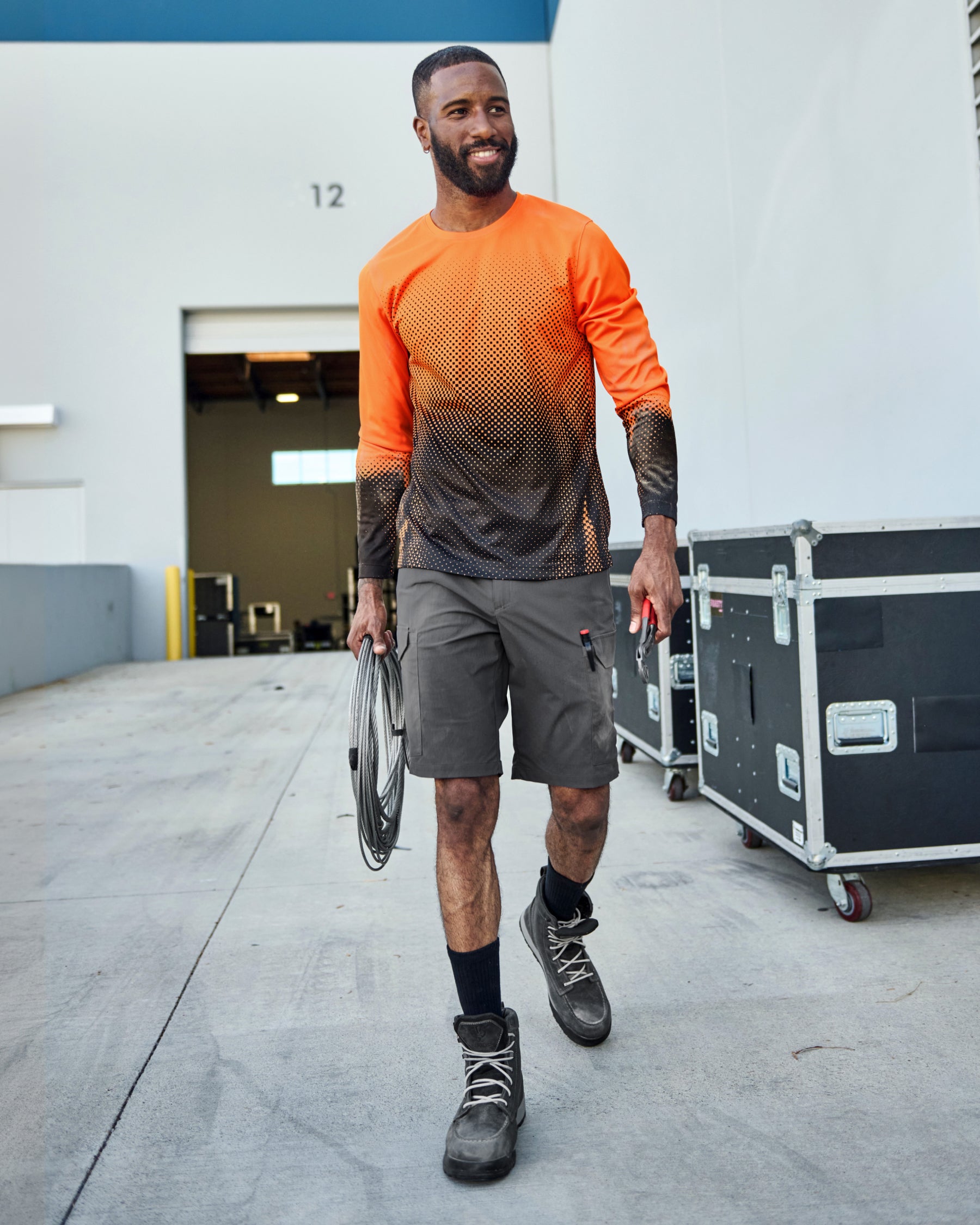 A worker wears the STRAUSS Enhanced Visibility Performance Long Sleeve Shirt e.s.t:aktik in neon orange and black with black work pants near equipment on a paved jobsite