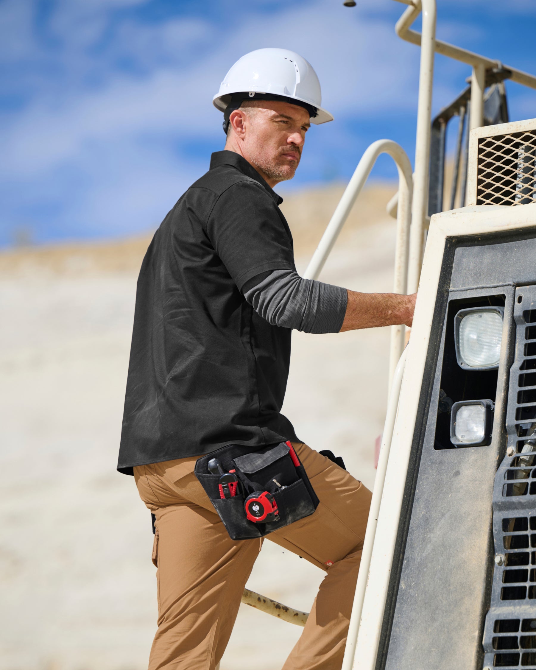 A worker wears the STRAUSS Short Sleeve Work Shirt e.s.iconic in black open over a T-shirt with cargo work pants near a worksite