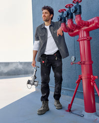 A worker wears the STRAUSS Short Sleeve Work Shirt e.s.iconic in carbon gray open over a T-shirt with cargo work pants near a worksite