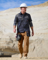 A worker wears the STRAUSS Short Sleeve Work Shirt e.s.iconic in nero blue open over a T-shirt with cargo work pants near a worksite