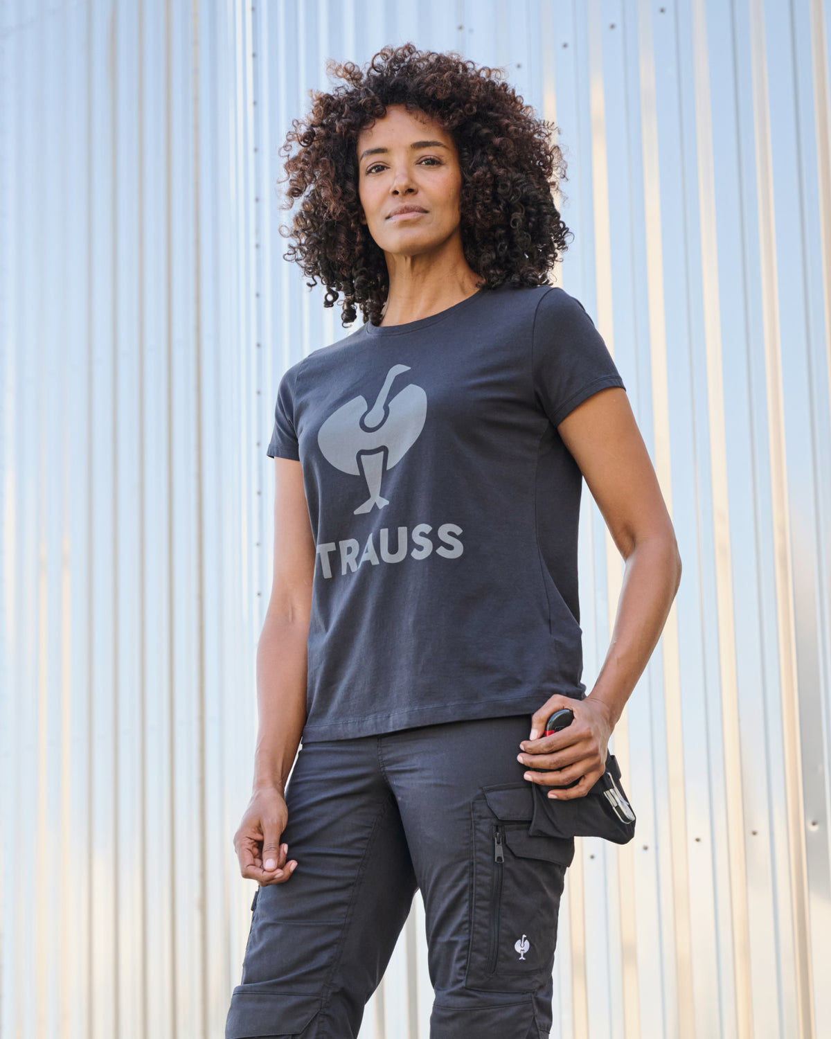 A woman wears the Women's STRAUSS T-Shirt e.s.concrete in black with work pants near a concrete wall with STRAUSS branding visible