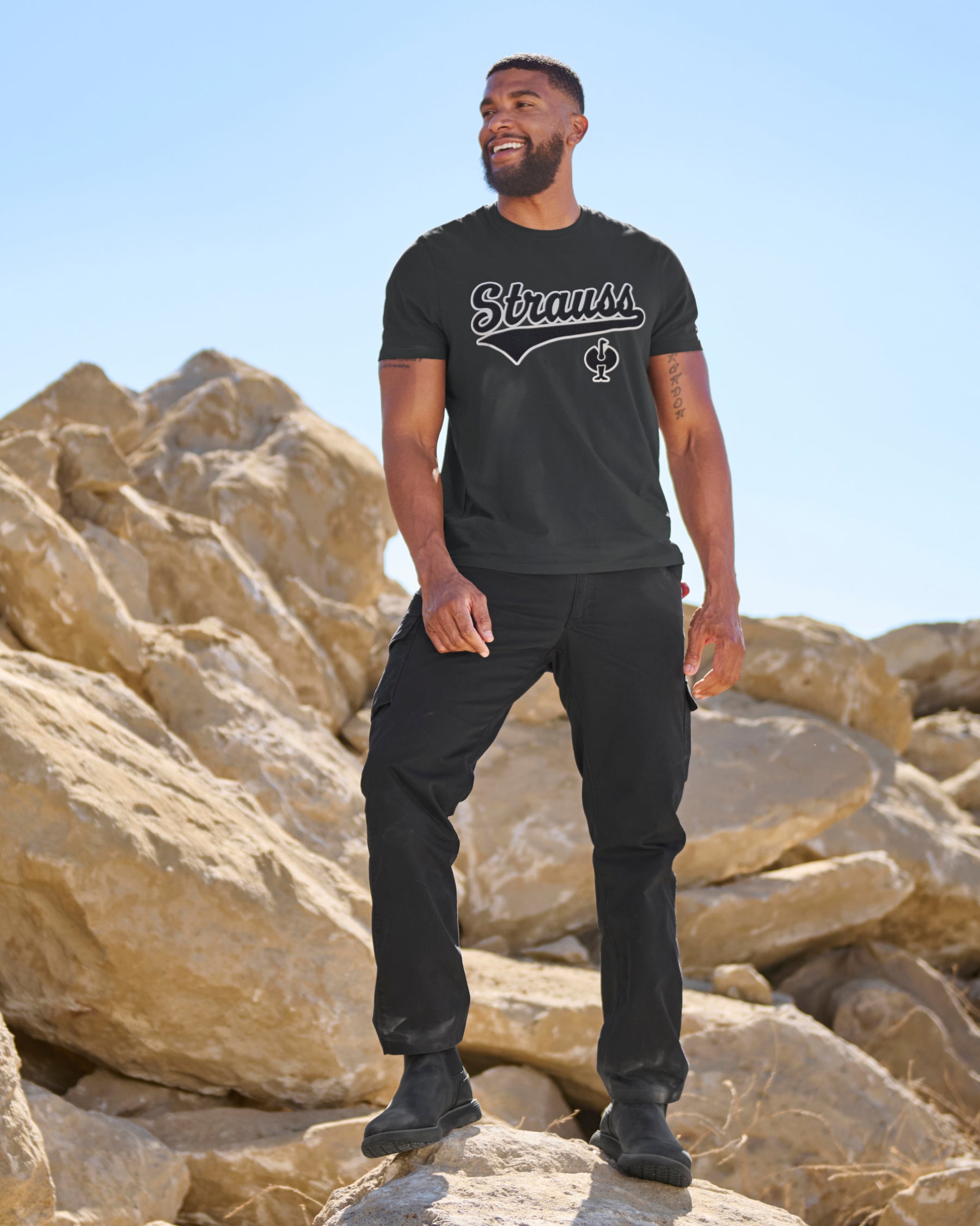 A model wears the STRAUSS Artwork T-Shirt e.s.e:pic in black with work pants while climbing over rocks