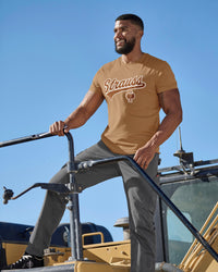 A model wears the STRAUSS Artwork T-Shirt e.s.e:pic in almond brown with work pants while climbing over rocks