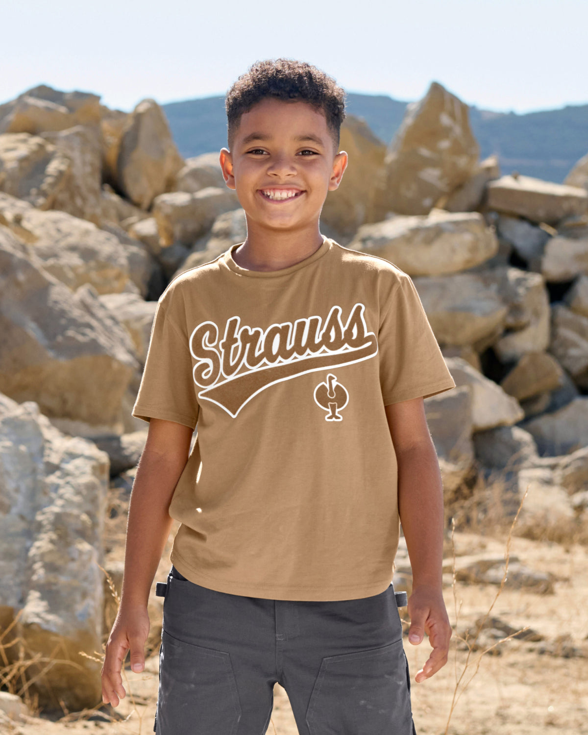 A child wears the Kids' STRAUSS Artwork T-Shirt e.s.e:pic in almond brown under a clear blue sky