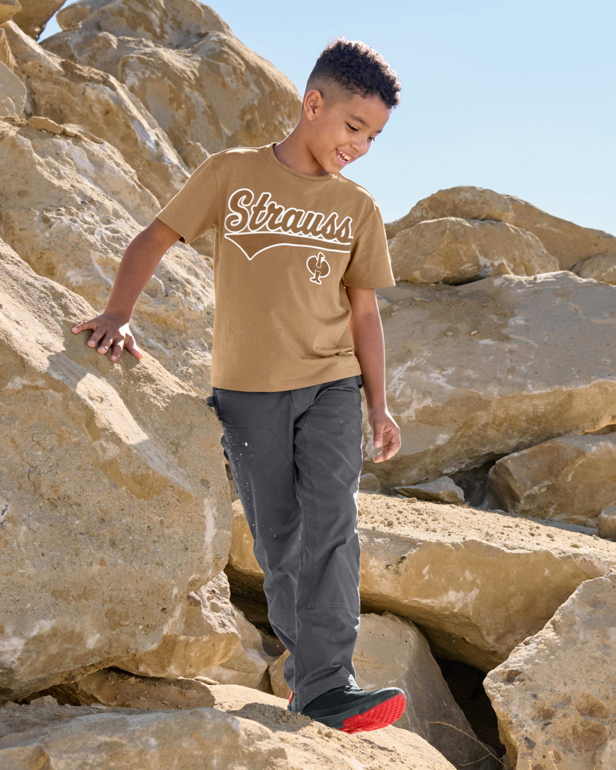 A child wears the Kids' STRAUSS Artwork T-Shirt e.s.e:pic in almond brown while holding a rock in a dry landscape