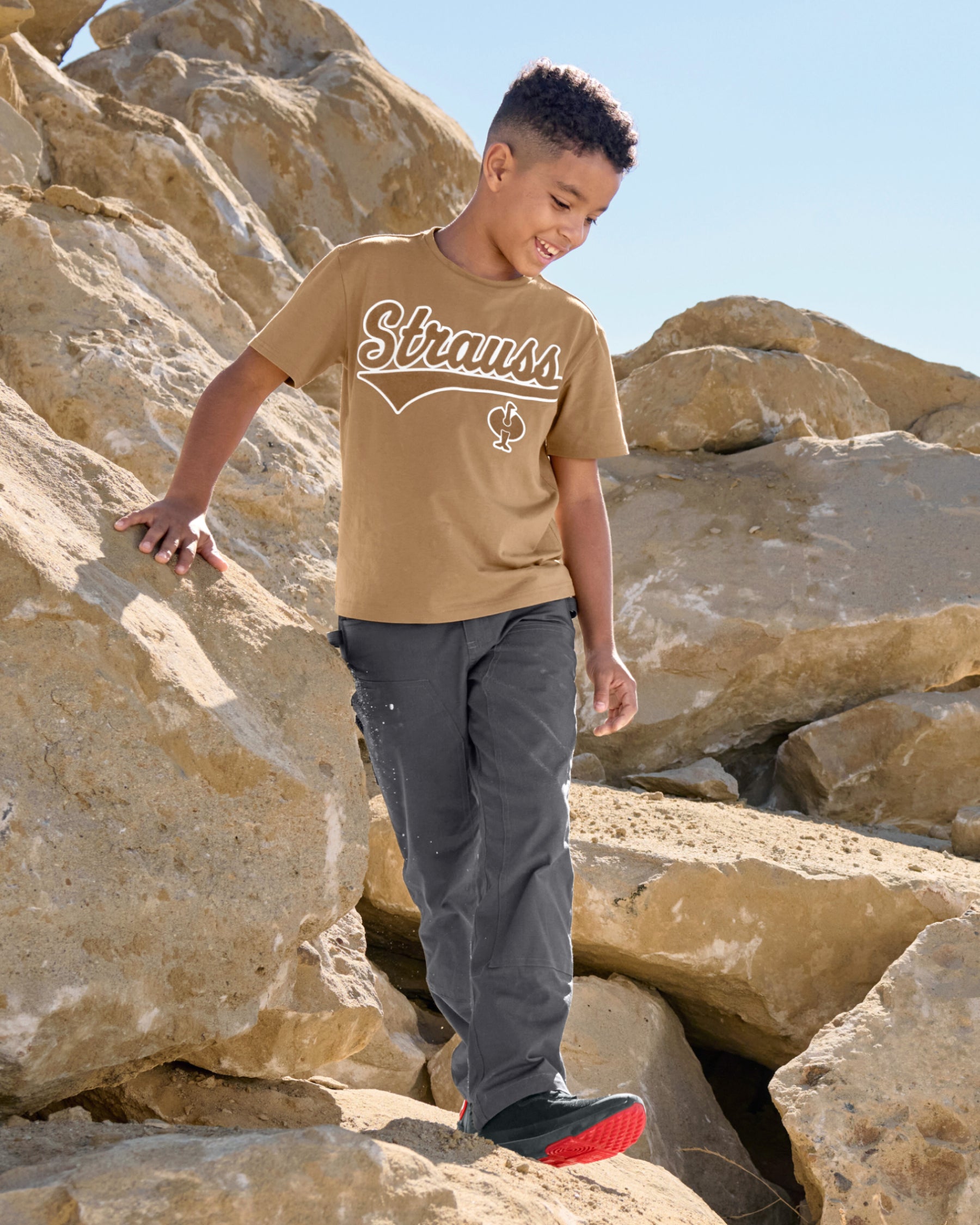 A child wears the Kids' STRAUSS Artwork T-Shirt e.s.e:pic in almond brown while holding a rock in a dry landscape