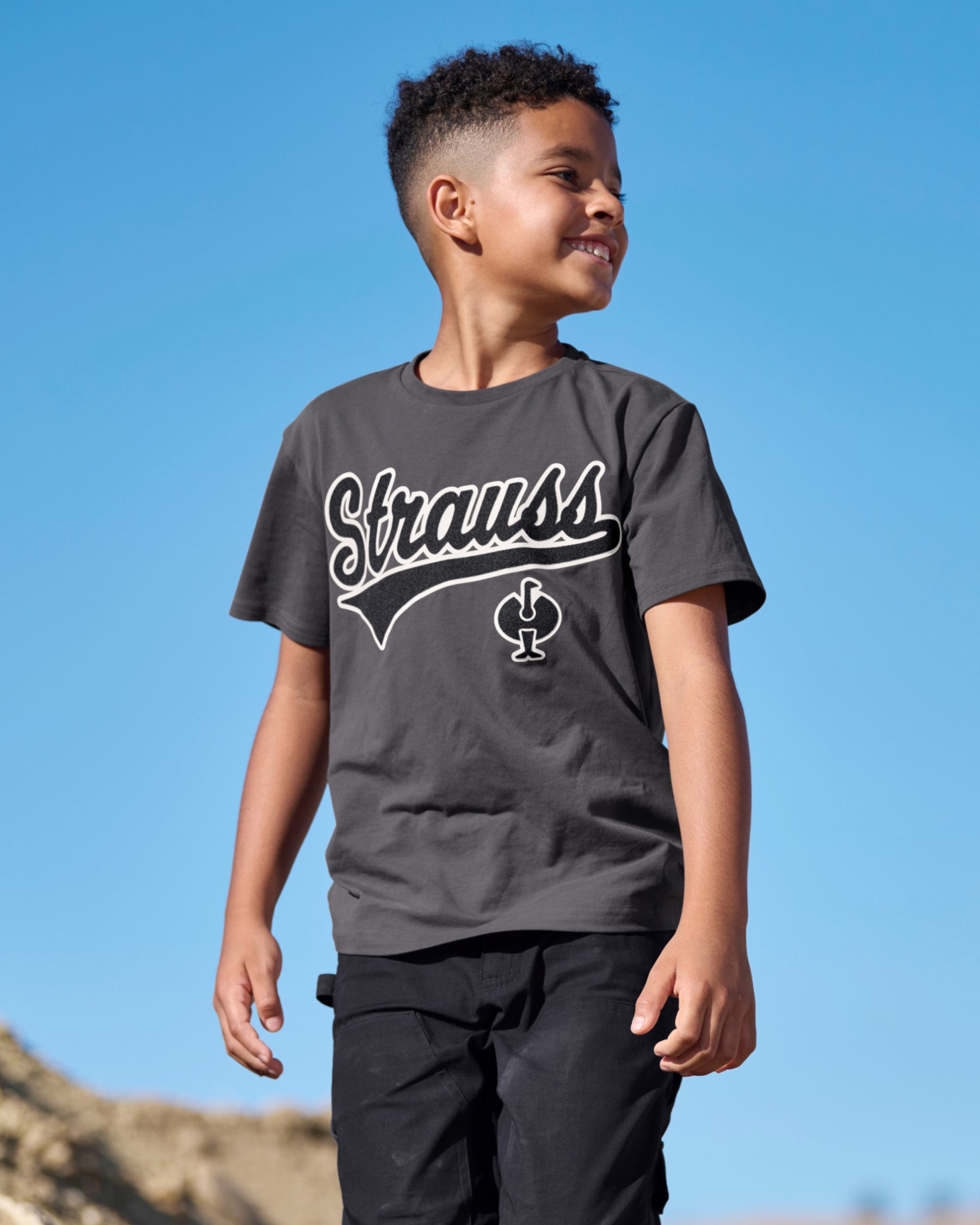 A child wears the Kids' STRAUSS Artwork T-Shirt e.s.e:pic in carbon gray under a clear blue sky