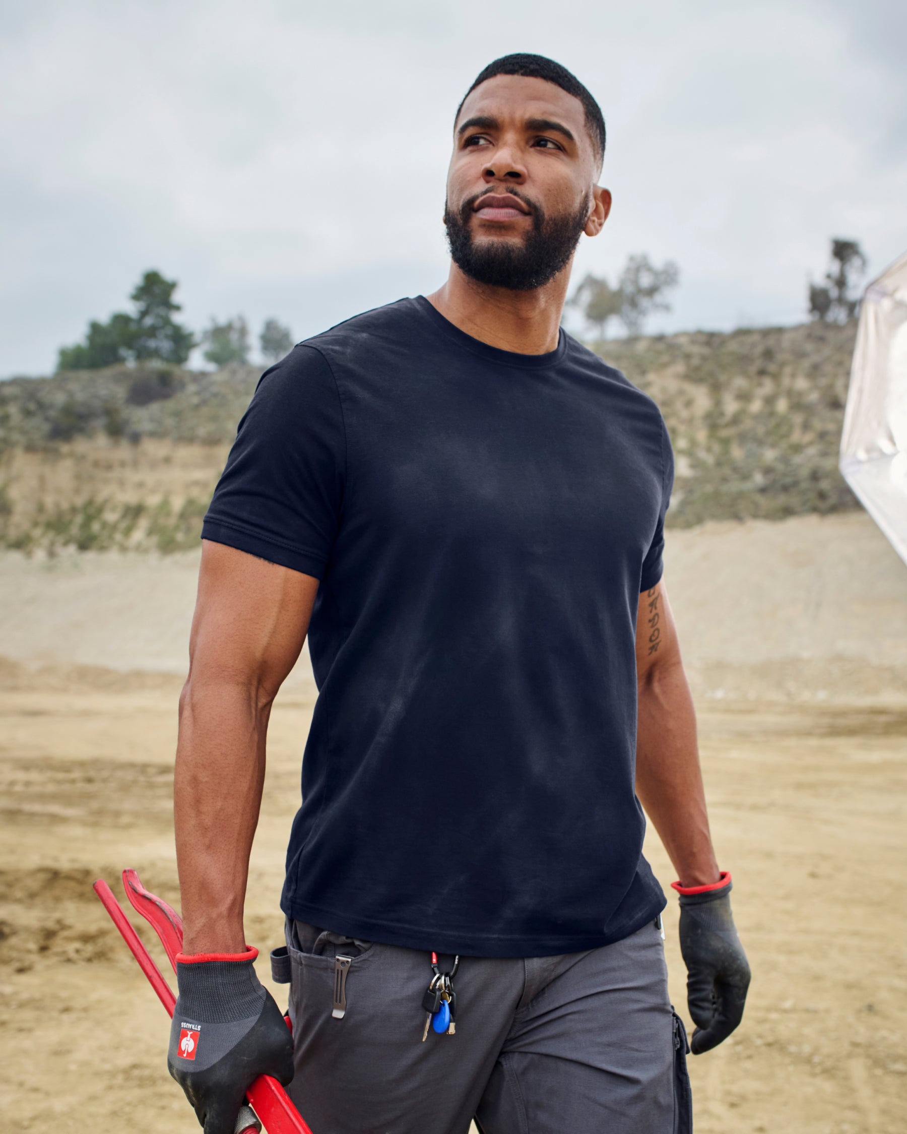 A worker wears the STRAUSS Cotton Stretch T-Shirt in navy with tan work pants beside jobsite equipment
