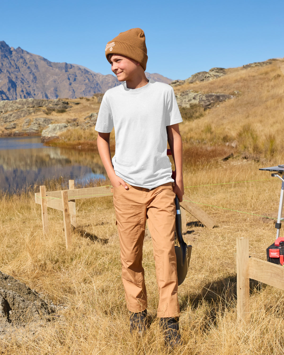A child wears the Kids' STRAUSS Cotton Stretch T-Shirt in white with kids' work pants while walking through a sunny field