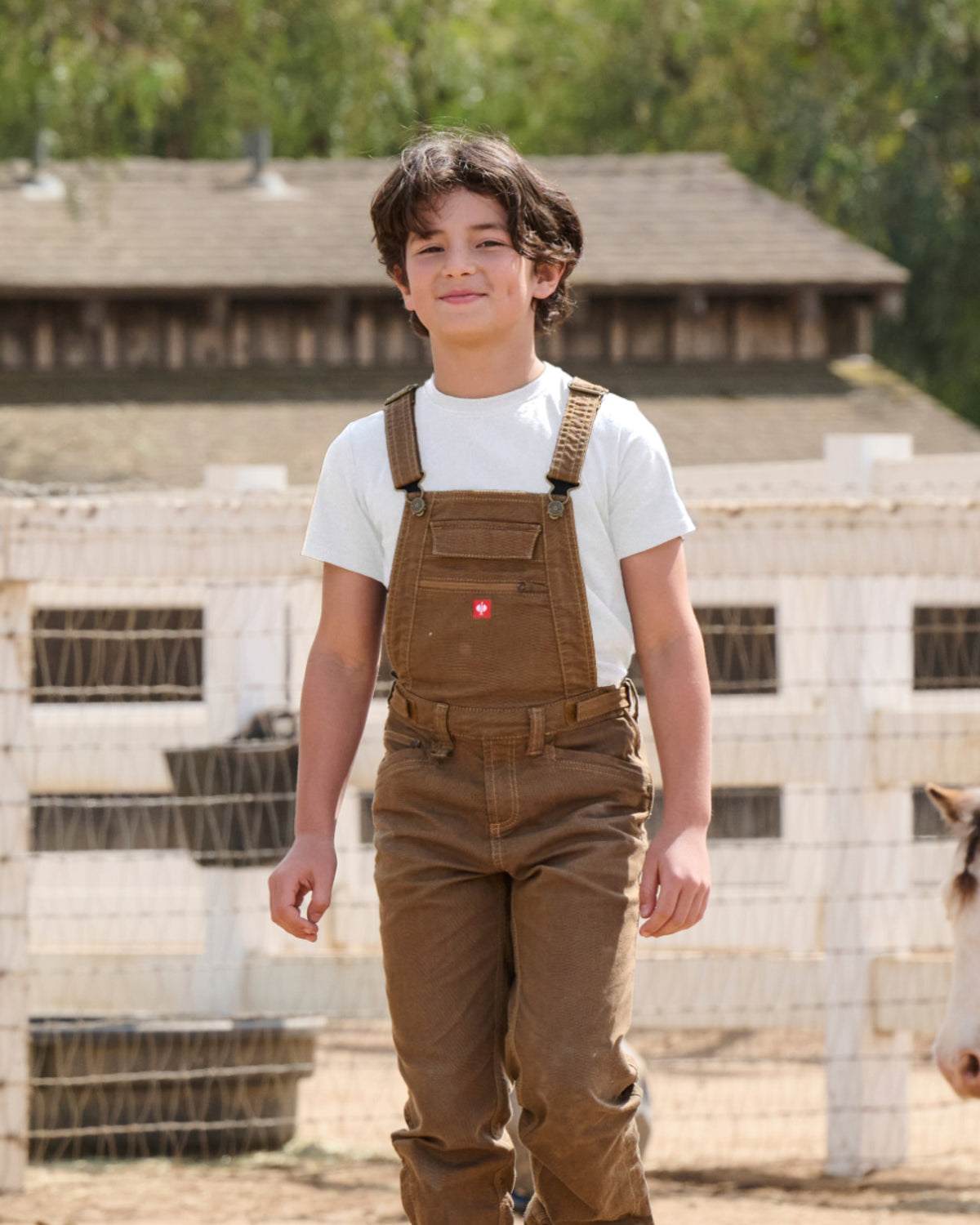 A child wears the Kids' STRAUSS Cotton Stretch T-Shirt in white with kids' work pants beside outdoor work gear