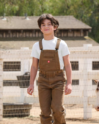 A child wears the Kids' STRAUSS Cotton Stretch T-Shirt in white with kids' work pants beside outdoor work gear