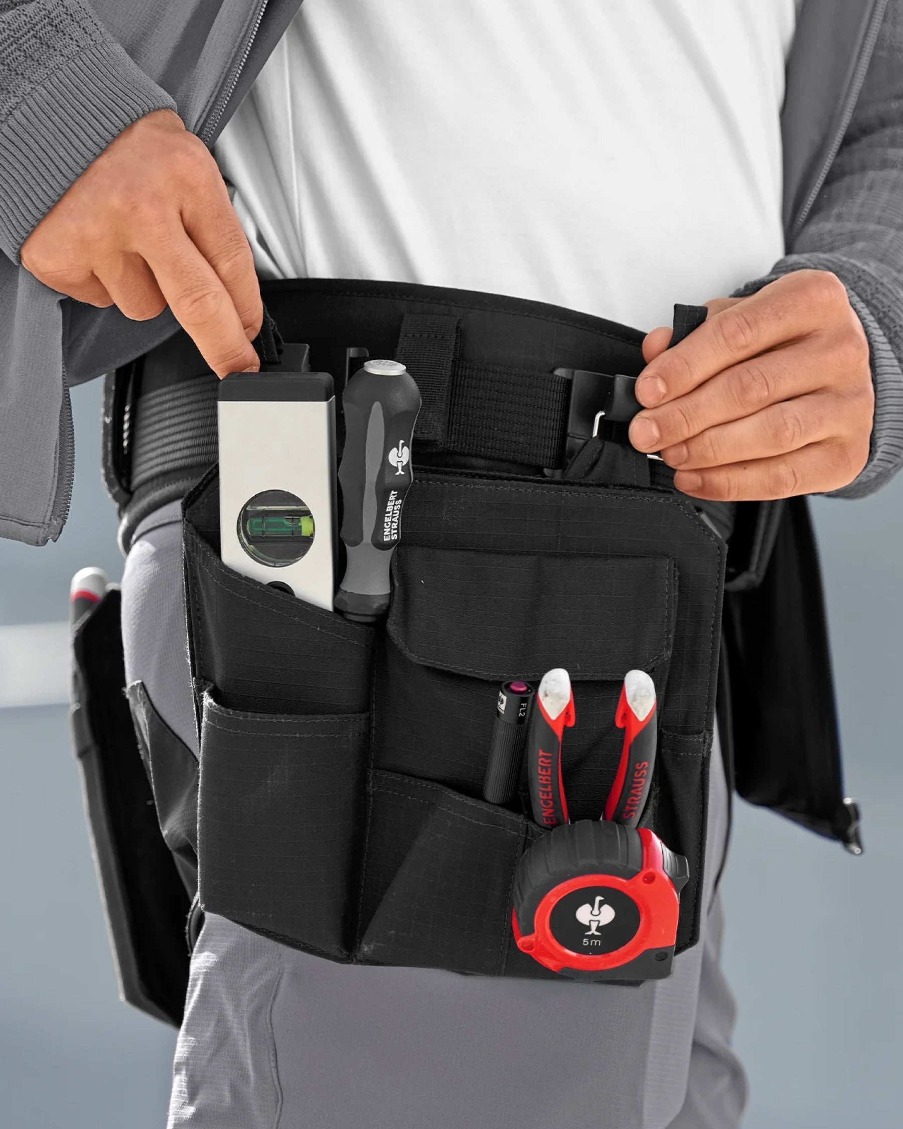 A black STRAUSS e.s.tool concept right-hand large tool bag is fastened to work pants while a worker reaches for hand tools.
