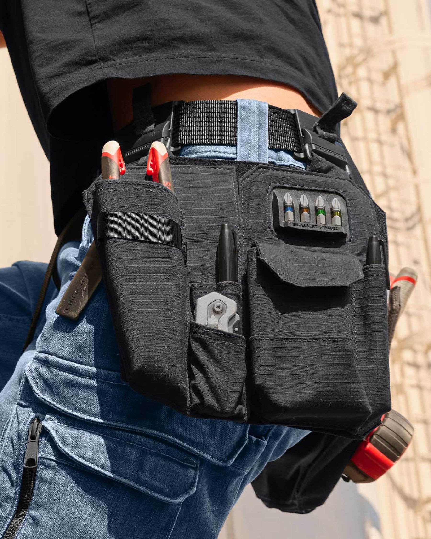 A black STRAUSS e.s.tool concept nails and screws bag sits on a work belt over utility pants in a jobsite scene.