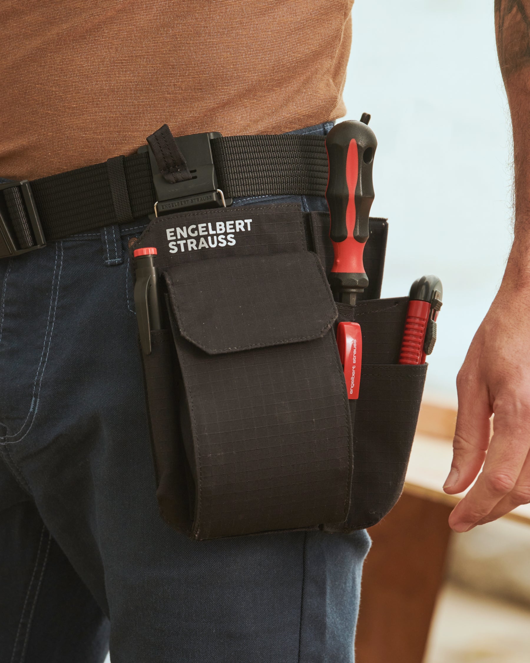 A black STRAUSS e.s.tool concept multipocket is fastened to work pants while a worker reaches for hand tools.
