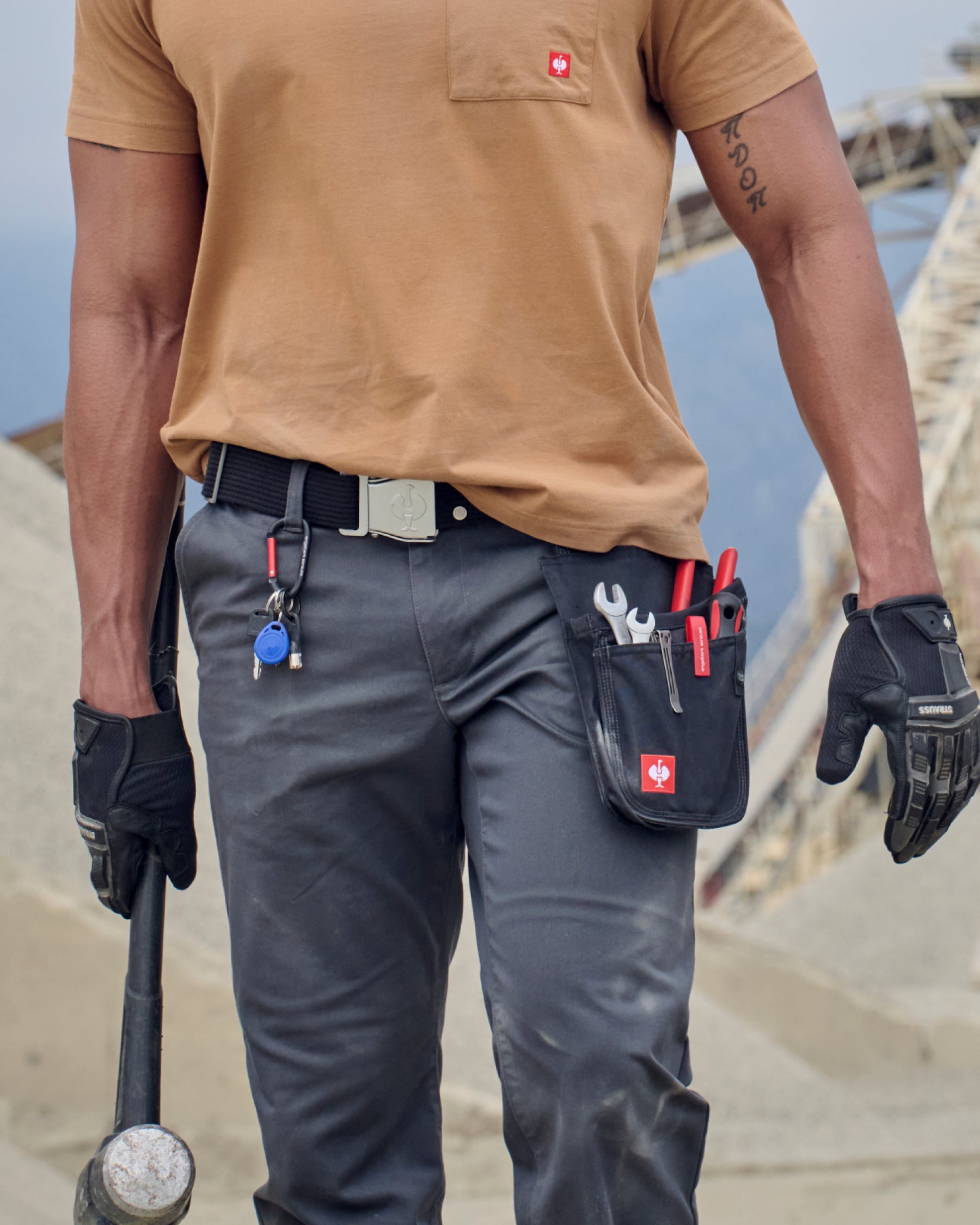 A black STRAUSS tool bag is fastened to work pants while a worker reaches for hand tools.
