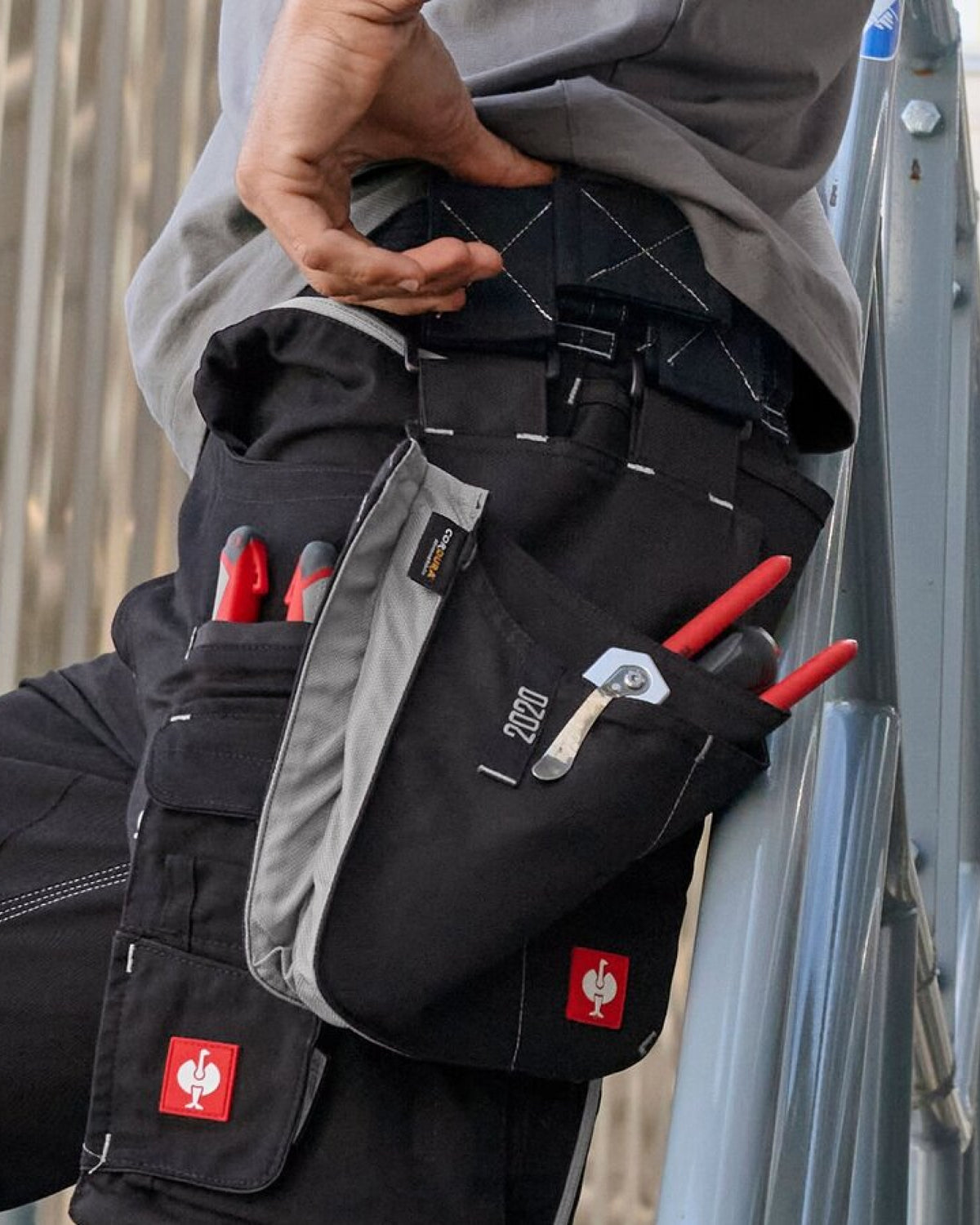 A black and platinum STRAUSS e.s.motion 2020 large tool bag is fastened to work pants while a worker reaches for hand tools.