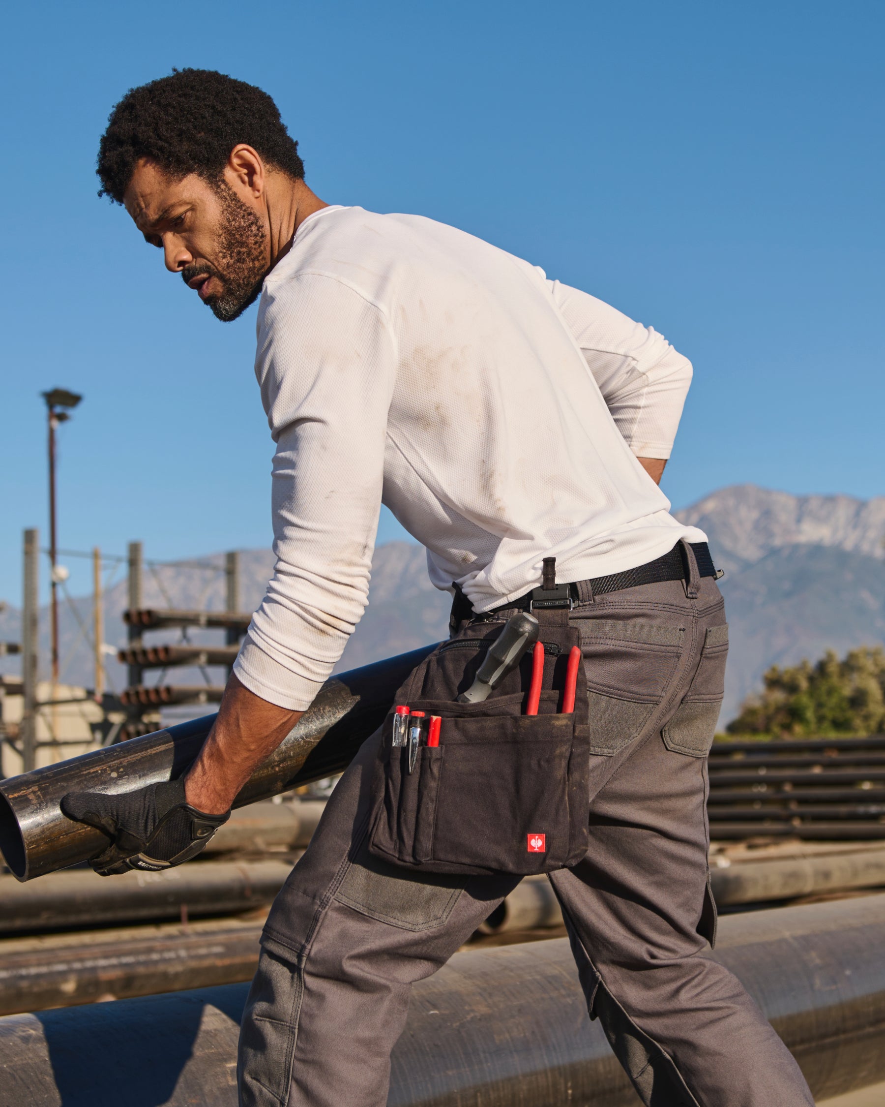 A black STRAUSS e.s.iconic tool bag is fastened to work pants while a worker reaches for hand tools.
