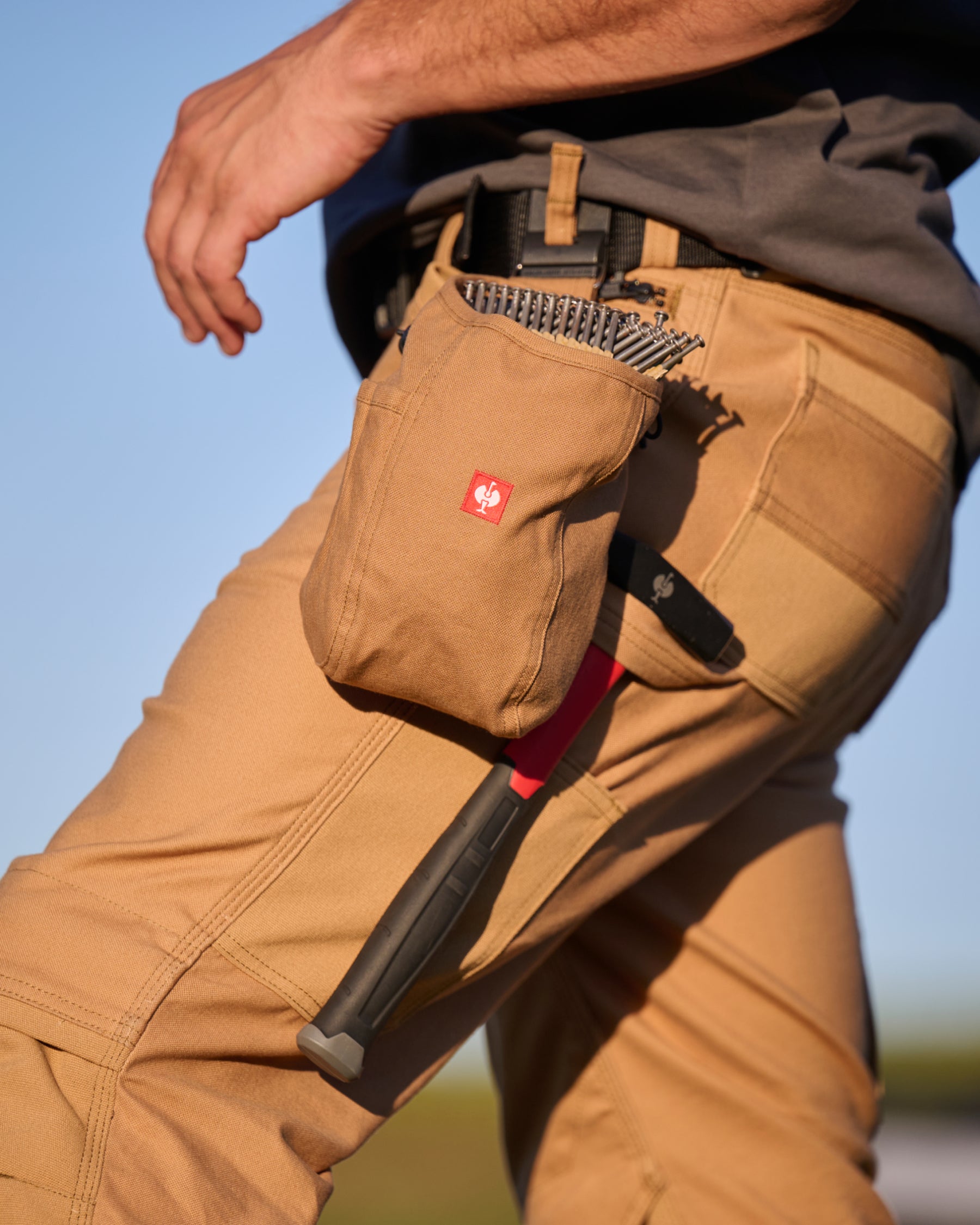 A almond brown STRAUSS e.s.iconic nail bag sits on a work belt over utility pants in a jobsite scene.